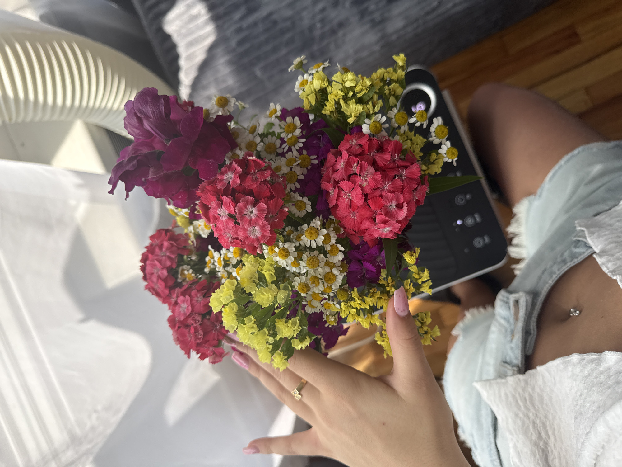 A close-up shot shows a person's hand, adorned with a gold ring and pastel pink nail polish, gently touching a vibrant bouquet of flowers. The bouquet is a colorful mix of red, pink, yellow, and white blossoms, with some delicate daisy-like flowers and deeper purple blooms. The hand is positioned in the lower right quadrant of the image, with the flowers filling the left and center.
In the background, the setting appears to be indoors, likely in a home. A light-colored, ribbed object, possibly a fan or air purifier, is visible on the left. To the right of the flowers, a dark electronic device with buttons is partially visible, suggesting a table or desk. Further back, a person's legs are visible, clad in light blue denim shorts, indicating they are sitting. The lighting suggests it is daytime, with sunlight casting shadows on the white surface to the left. The overall mood is bright and cheerful, emphasized by the colorful flowers. No text is visible in the image.