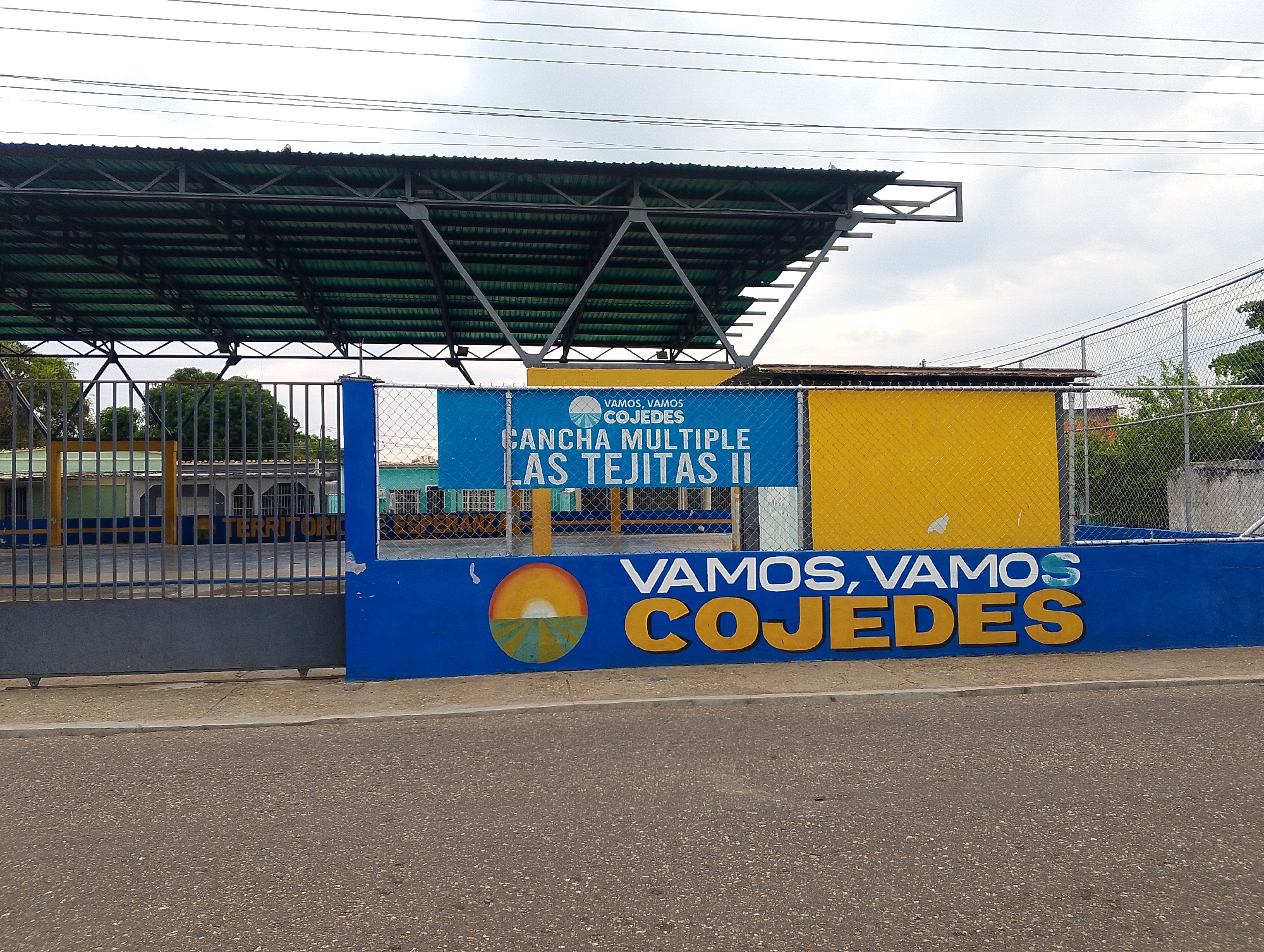 The image depicts a street scene in San Carlos, Venezuela, featuring the exterior of a sports facility. A blue wall, emblazoned with the words "VAMOS, VAMOS COJEDES" in yellow, runs along the front of the facility. A circular logo featuring a stylized depiction of a sunrise over a field is adjacent to the text. Behind the wall, a chain-link fence encloses the facility. A blue banner that says “VAMOS, VAMOS COJEDES”, “CANCHA MULTIPLE LAS TEJITAS II” hangs on the fence. A covered structure with a green roof and a metal frame is visible within the facility. To the left, a partially visible gray metal gate stands in front of a building with green and yellow details. Overhead, power lines stretch across the sky. The foreground consists of a paved road.