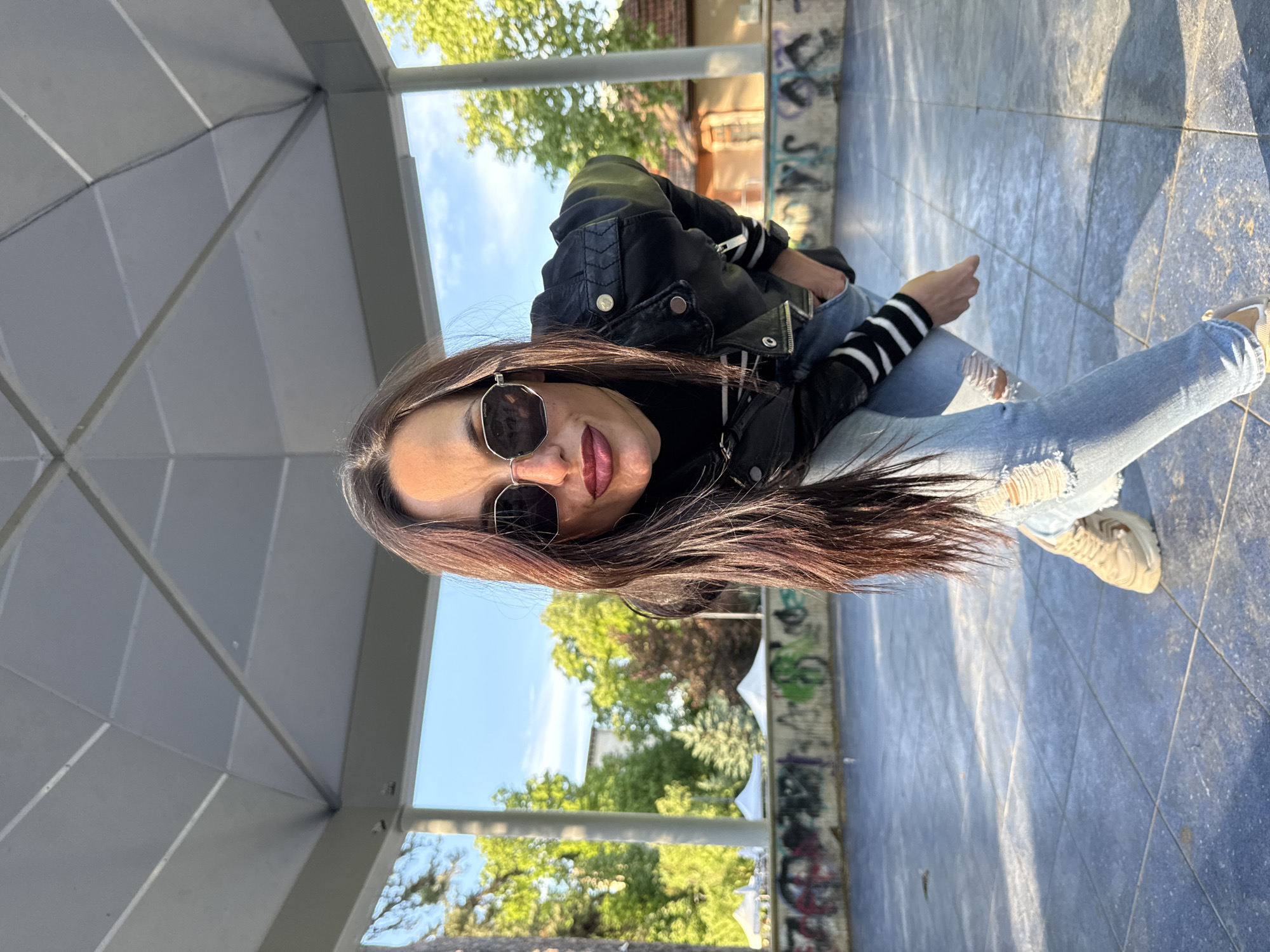 A woman with long brown hair, wearing sunglasses, a black leather jacket with striped sleeves, and ripped jeans, is sitting under a gazebo-like structure. She is facing the camera with a slight smile. The gazebo has a grey roof and is supported by pillars. Outside the gazebo, there is greenery, including trees and bushes. In the background, there is graffiti on a wall. The ground is tiled with blue and grey tiles. The location is likely in the city of Pleven, Bulgaria.