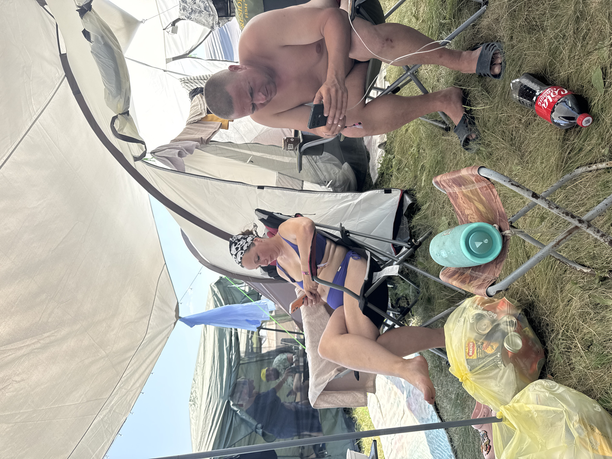 In a grassy outdoor area, two adults are seated in folding chairs under a large tent. The tent is white and appears to be set up for camping, with other tents visible in the background. The sky is clear and blue.
On the left, a woman wearing a purple bikini and a black and white bandana is seated, looking at her orange smartphone. She has long, tan legs and is sitting in a black folding chair. To her left, a yellow plastic bag filled with cans and bottles is on the ground.
On the right, a shirtless man with short hair is seated, also looking at his black smartphone. He is wearing dark flip-flops. To his right, a bottle of Coca-Cola is lying on the grass. A turquoise speaker is resting on another folding chair in the foreground.
In the background, other campers are visible, including one person in a green shirt wearing a yellow hat, who is also looking at a phone. Clothing hangs from a line within the tent. The overall scene suggests a casual outdoor gathering or camping event.