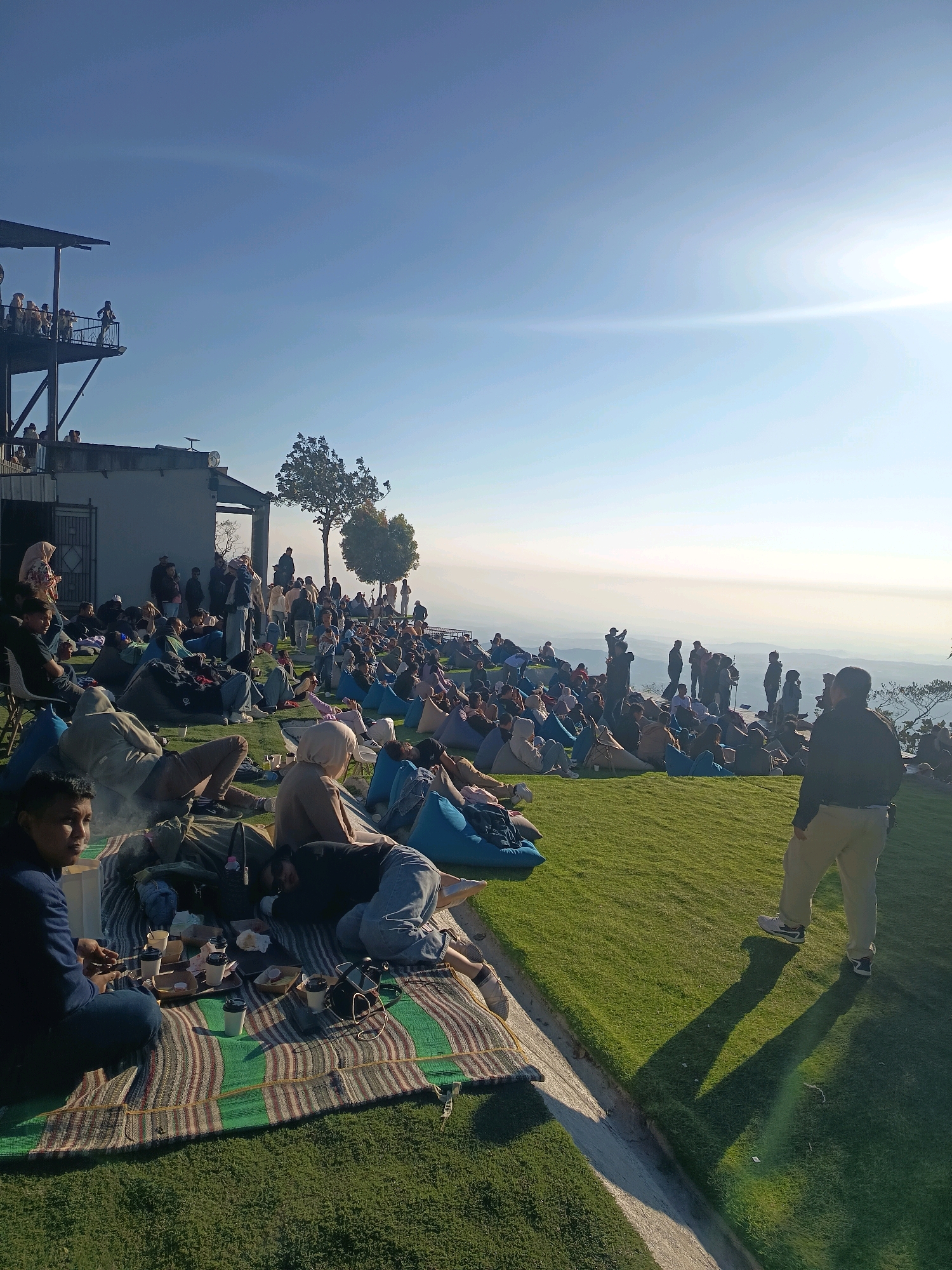 In Keditan, Indonesia, a large group of people are gathered on a grassy hillside for what appears to be an outdoor event. The setting sun casts long shadows across the lush green lawn, and the sky is a clear, bright blue with a sun flare visible on the right. Many individuals are seated on colorful beanbag chairs or picnic blankets, some with food and drinks spread out before them. A man in the foreground, facing away from the camera, stands on the grass. In the background, a multi-level structure with a balcony overlooks the scene, with several people visible on the balcony. The landscape stretches out towards a hazy horizon, suggesting a high vantage point.