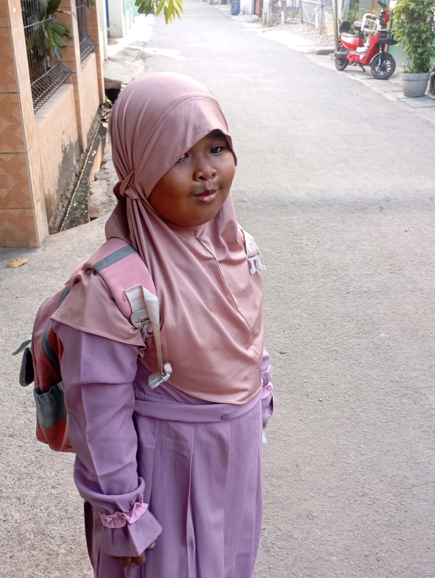 The image shows a young girl in a pink hijab and a purple dress with a backpack, standing on a paved street in Babelan, Indonesia. She is facing right, with her head slightly turned towards the camera. The background shows a building with orange tiles, a drain, a fence, and a red scooter parked on the side of the road. Trees and a potted plant are also visible. The lighting suggests it is daytime.