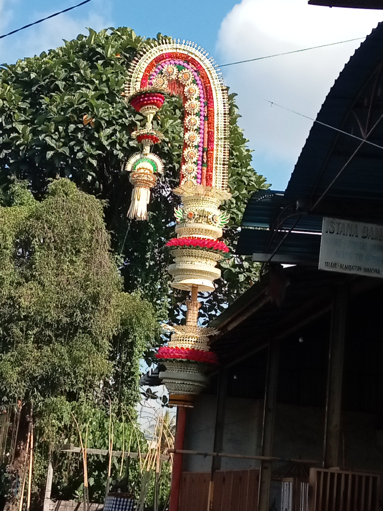 A towering, ornate, and vibrantly colored decorative structure, possibly a Balinese temple offering called a Canang Sari, is the central focus of this outdoor scene. The structure is a tiered arrangement of woven materials, intricate carvings, and bright floral elements, culminating in a tall, arched top adorned with pink, yellow, and green accents. It stands against a backdrop of lush green trees and a bright blue sky with scattered white clouds. To the right of the decorative structure, a dark, sloped roof of a building is visible, with a sign that reads "ISTANA BANA" followed by some less legible text. The building appears to be made of dark wood and metal. The foreground shows more greenery and some wooden fence-like structures, suggesting an outdoor market or a residential area. The overall impression is of a festive or religious occasion in a tropical environment.