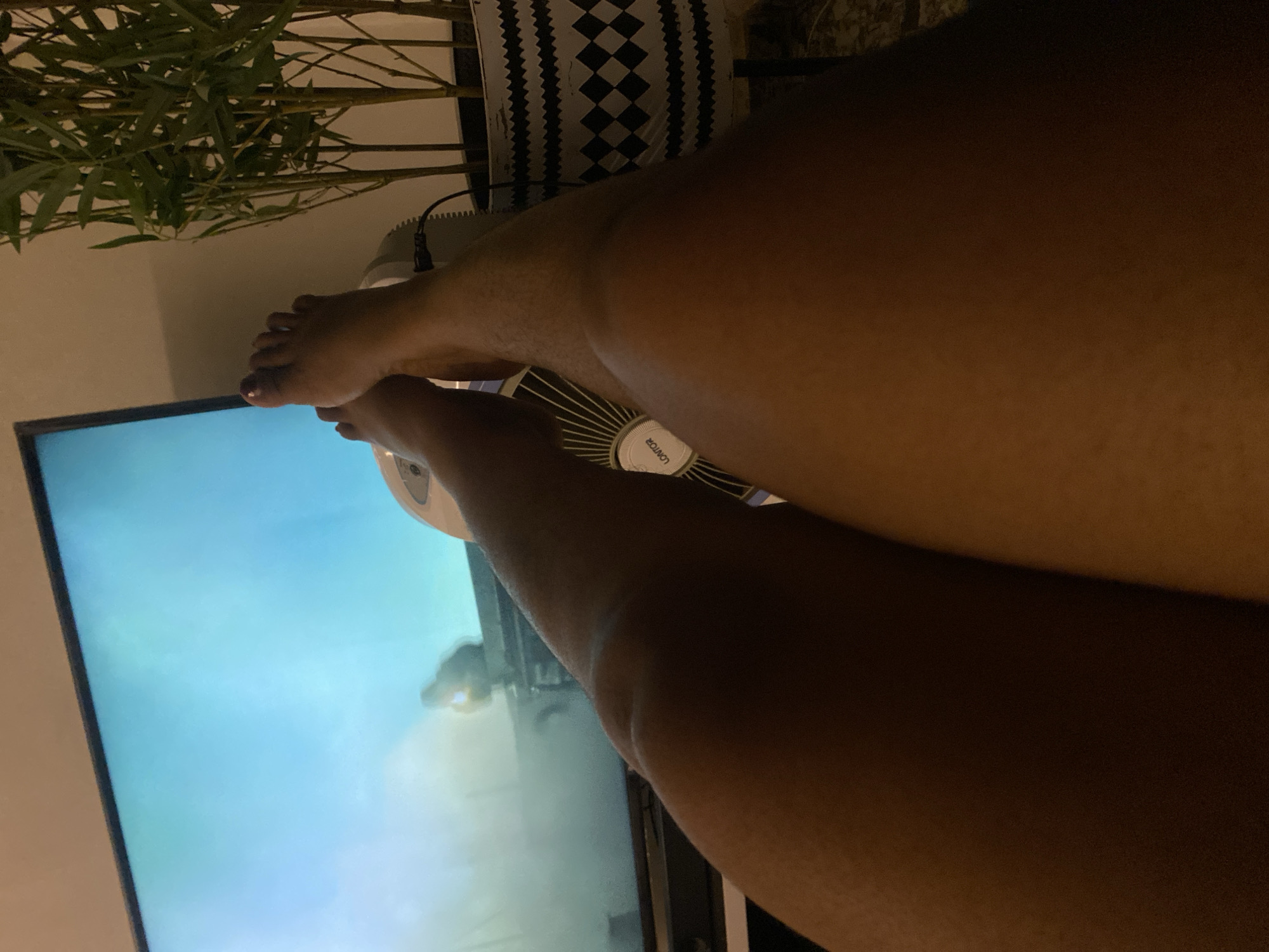 The image shows a person's legs resting on a white fan in front of a television. A house plant sits behind the fan. The context of Abuja, Nigeria, cannot be determined from the image.