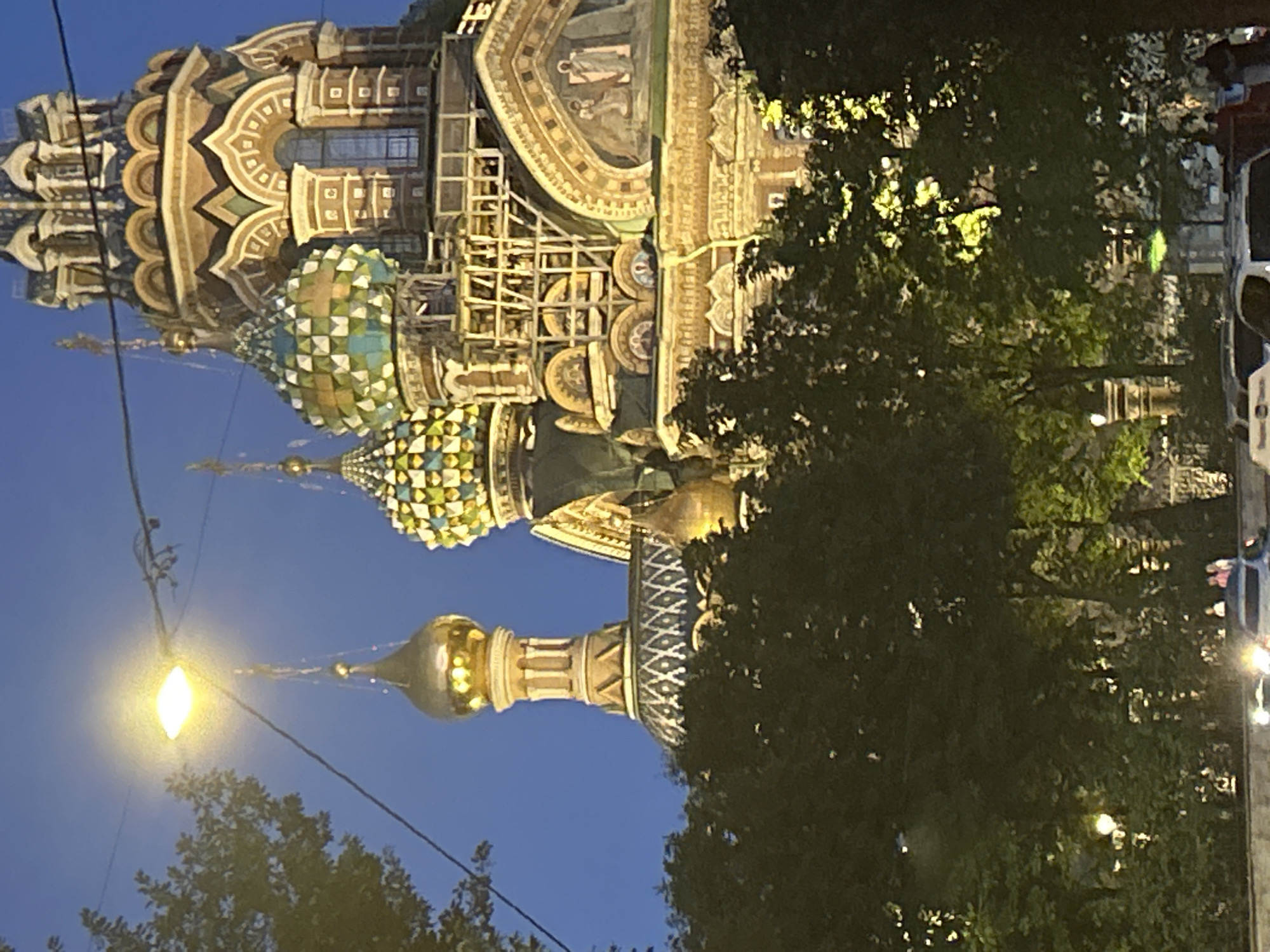 In the city of Unknown, Unknown, a street view at dusk captures the ornate, multi-domed Church of the Savior on Spilled Blood. The sky is a deep twilight blue, punctuated by the bright glow of a streetlamp and overhead wires. The church's distinctive onion domes, one adorned with gold and others with colorful mosaics of green and yellow, rise majestically. The building's facade features intricate architectural details, including arched windows and decorative patterns, with scaffolding suggesting ongoing construction or renovation. The lower portion of the image is dominated by dark green foliage, likely trees, partially obscuring the church and the street below. In the foreground, the headlights of parked cars are visible, and a few individuals can be seen in the distance, suggesting the presence of pedestrian activity near the church.