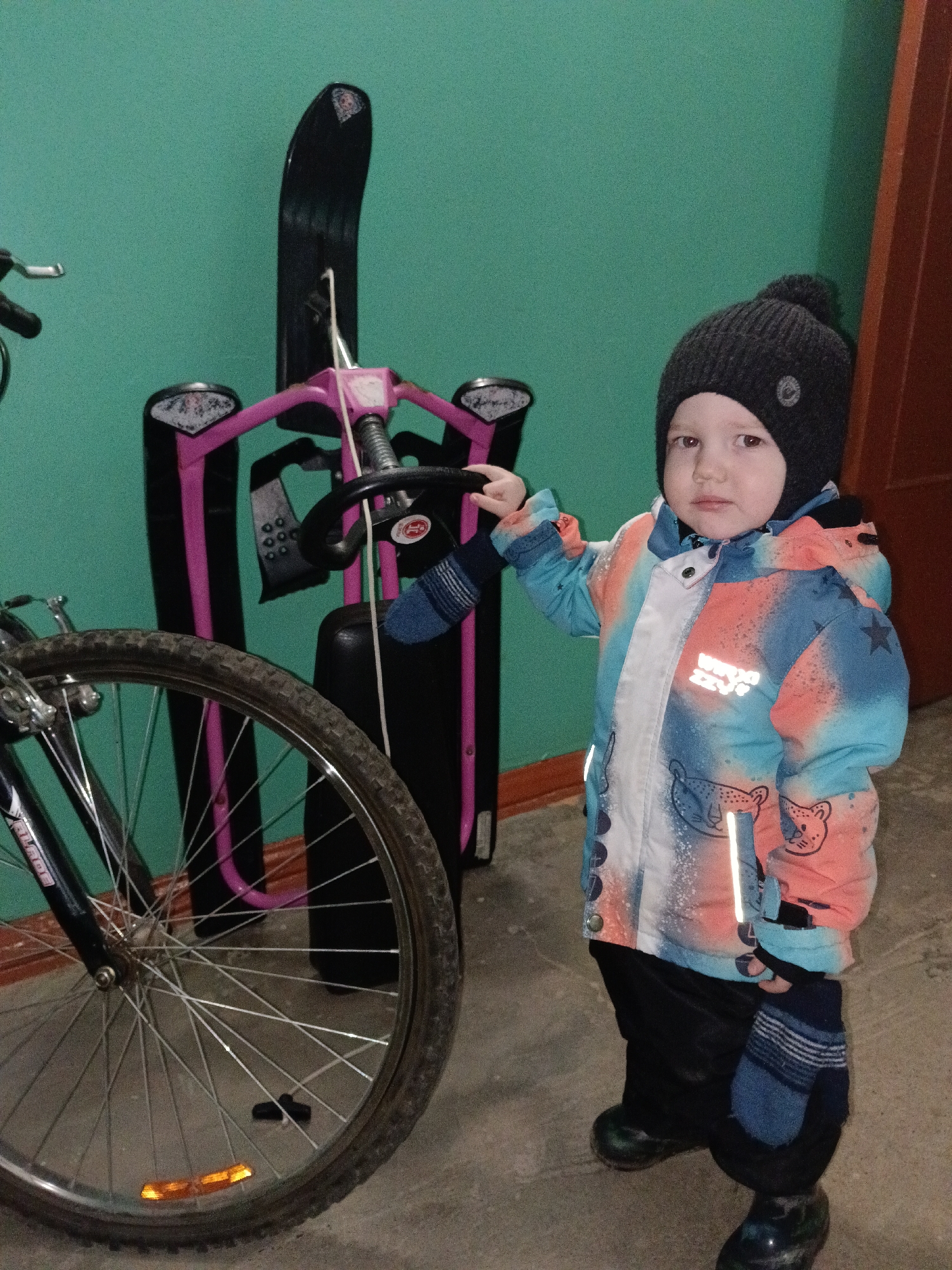 The image captures an indoor scene in Chaikovsky, Russia. A young child, likely a toddler, stands in the center-right of the frame, facing forward with a neutral expression. The child wears a dark grey knitted beanie with a pom-pom, a multi-colored winter jacket (blue, orange, white) adorned with animal prints and reflective strips, dark pants, and dark boots. Blue and white striped mittens cover both hands. The child's right hand rests on the black steering wheel of a snow sled, which features a prominent pink frame and a black body/seat, with a white strap hanging from the steering mechanism. To the left, the front wheel of a dark-colored bicycle is visible, equipped with a black tire, silver spokes, and an orange spoke reflector. A black brake pad lies on the floor near the bicycle wheel. The background consists of a solid turquoise-colored wall, with a dark brown wooden door frame partially visible on the far right. The floor appears to be a light-colored concrete or worn tile surface.