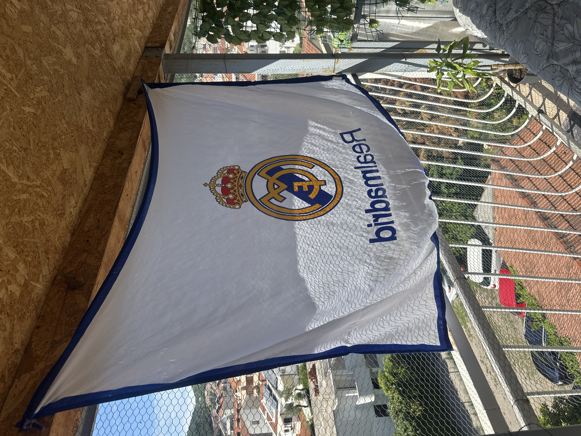 The image displays a Real Madrid football club flag hanging vertically on what appears to be an enclosed balcony or terrace. The white flag features the club's crest – a gold crown above intertwined blue and gold letters (likely "MCF" for Madrid Club de Fútbol) – and the word "Realmadrid" written in blue capital letters, which appears reversed due to the flag's orientation. The flag has a distinct blue border.
To the left, a wall is paneled with light brown Oriented Strand Board (OSB). The foreground and right side of the image are dominated by a white metal railing, behind which a dark mesh safety net is installed, suggesting a safety measure for the balcony. Green foliage, possibly artificial, lines the top of the railing, and a small potted plant is visible.
The scene is brightly lit by strong sunlight, casting clear shadows and indicating a sunny daytime. Beyond the railing, a residential landscape unfolds, characterized by buildings with distinctive red-tiled roofs, typical of Mediterranean architecture found in places like Budva, Montenegro. Green trees and other vegetation are interspersed among the structures. A road is visible with at least two parked cars, one white and another dark-colored (partially obscured), providing a glimpse into the street below. No people are visible.