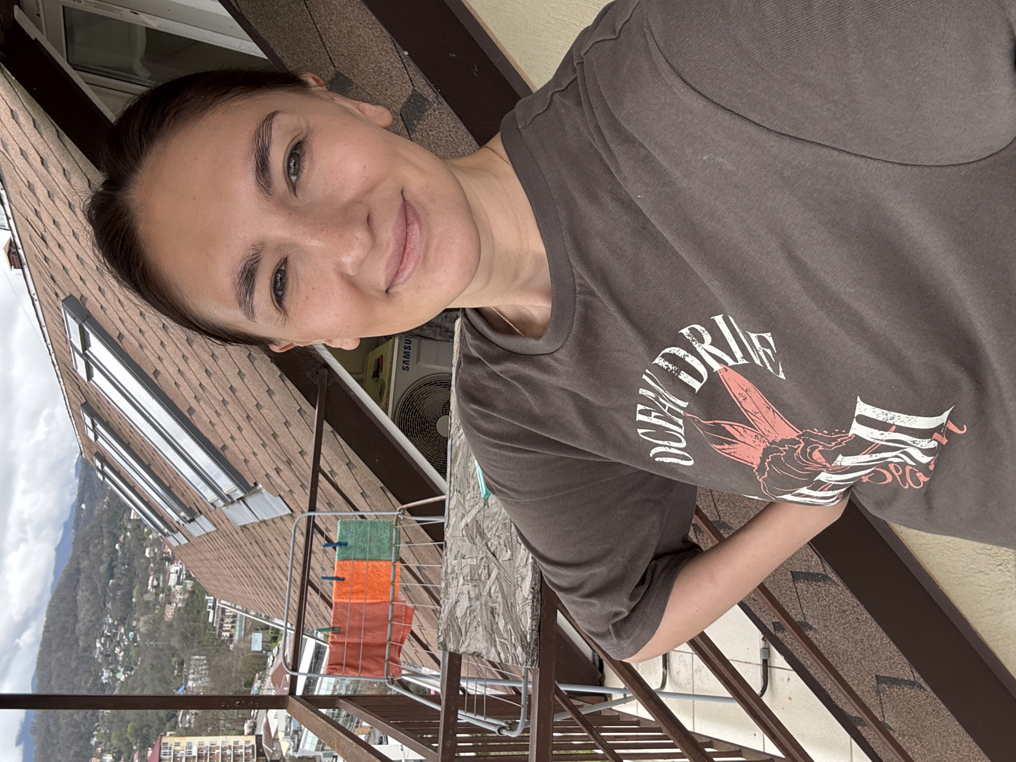 A woman in a brown t-shirt with a graphic design poses for a selfie on a balcony overlooking Sochi, Russia. Laundry hangs on a rack on the balcony. A Samsung air conditioner is visible in the background, mounted on the building. The landscape includes rooftops, distant mountains, and buildings.