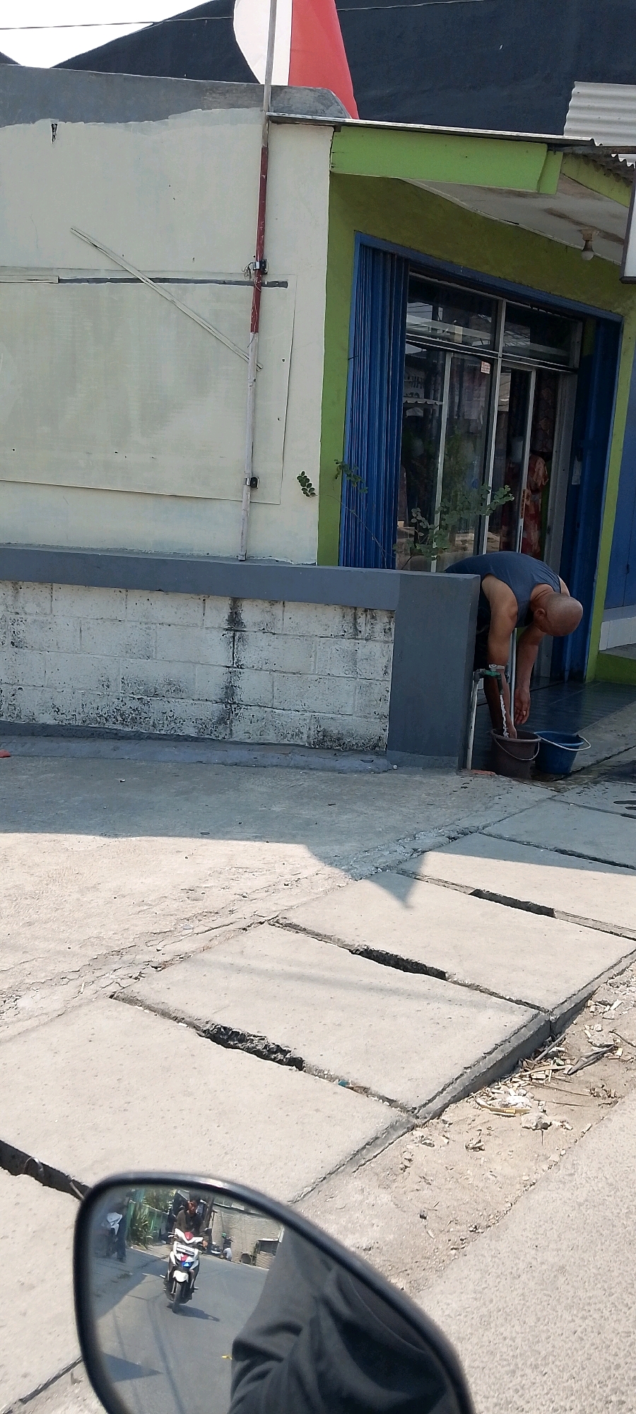 In Babelan, Indonesia, a bald man in a dark blue tank top is washing his hands in a bucket outside a building. The building has a light green and cream facade with blue metal folding doors. A white brick wall with some water stains runs along the front of the building. In the foreground, a cracked concrete sidewalk is visible. A motorcycle is reflected in a side mirror, showing a street scene with other motorcycles and people.