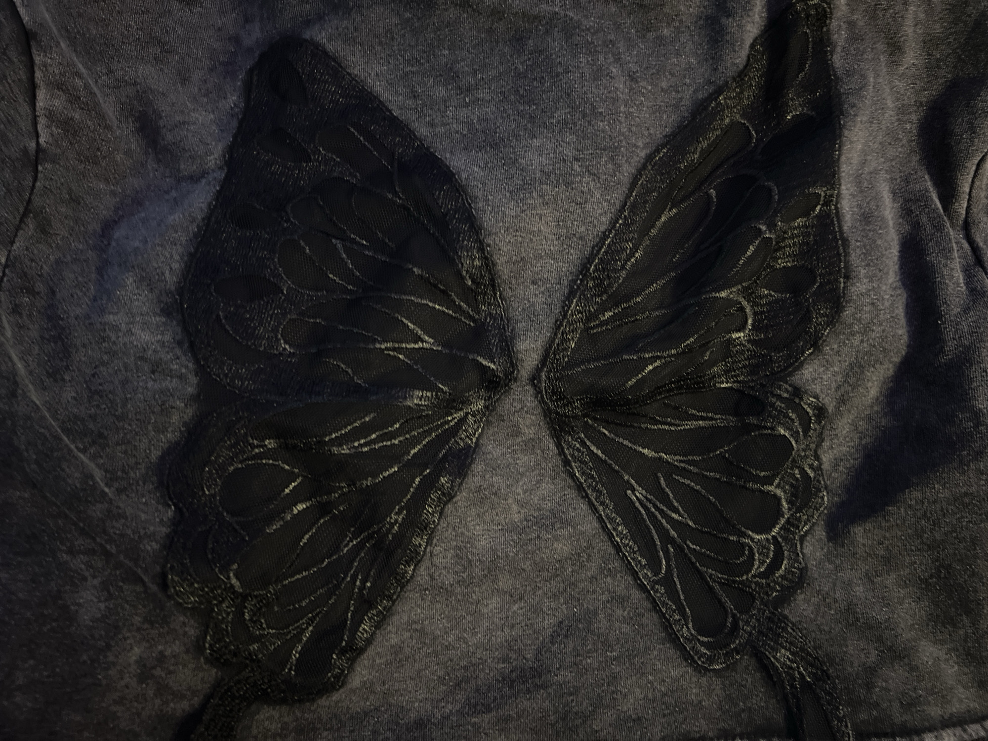 A close-up, overhead shot shows a dark gray fabric with a butterfly wing design. The design is in black lace and is split down the middle, creating two symmetrical wings. The wings are detailed with vein-like patterns and a scalloped edge. The fabric itself has a slightly textured, faded appearance, suggesting it could be a t-shirt or sweatshirt. The lighting casts shadows, particularly on the right side of the image, highlighting the texture of the fabric and the delicate nature of the lace. There is no visible text or indication of a specific location or time of day. The focus is entirely on the garment's design and material.