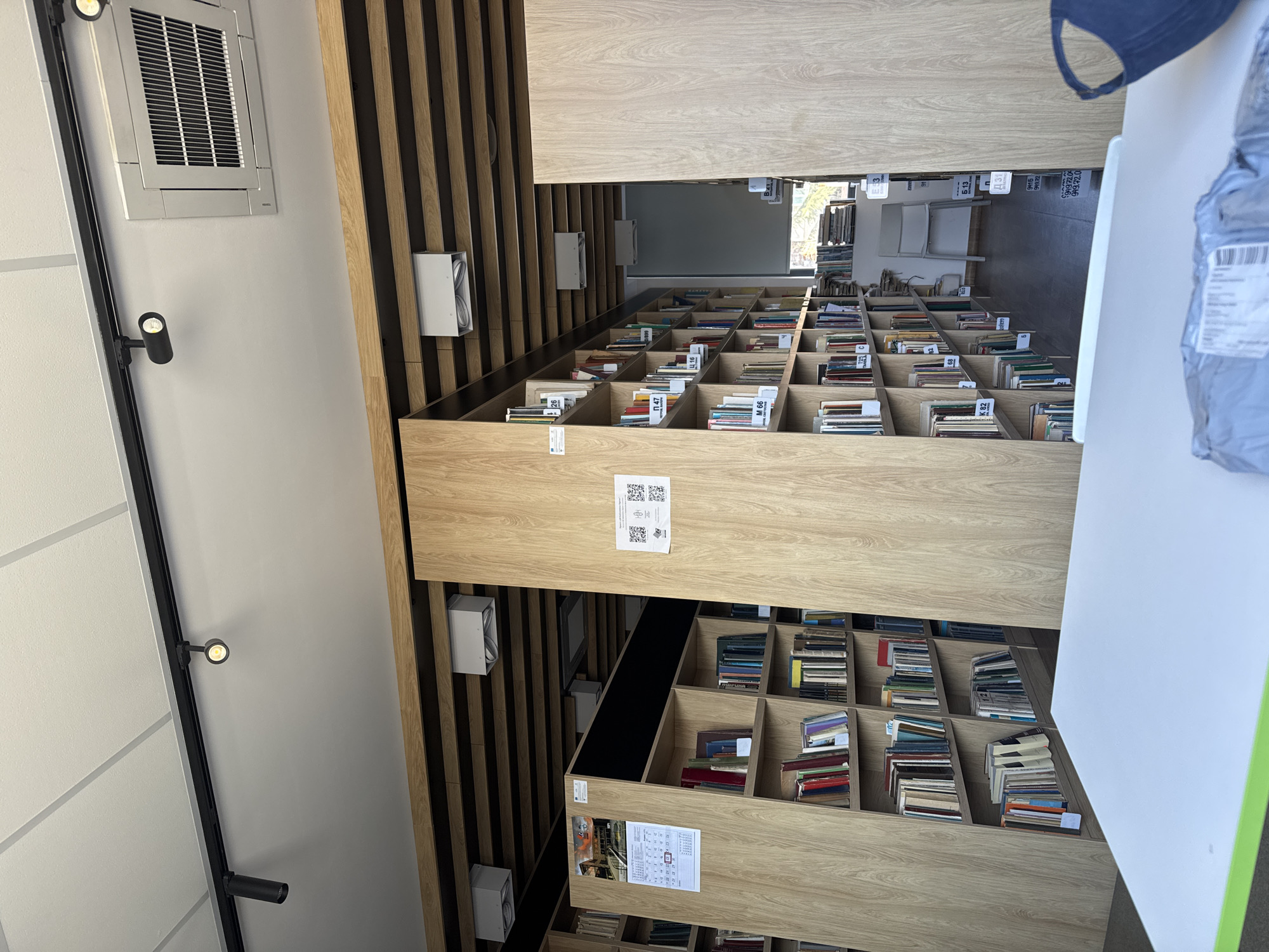 The image shows a bookshelf-lined hallway, seemingly in a library setting. The bookshelves are made of light-colored wood and are filled with books. Each section of the shelf has a label with what appears to be a call number on it. There is an air conditioning unit on the ceiling and a track lighting system along the wall. The books are arranged in neat rows, and the overall impression is one of order and organization. In the background, you can see chairs and what appears to be an outdoor setting.
The labels visible include "Π 47," "M 66," "Ц 16," "53," "Б 13," "K 82," "Д 31," and "949.72".
The library seems to be located in the city of Burgas, Bulgaria, as indicated by the location context.