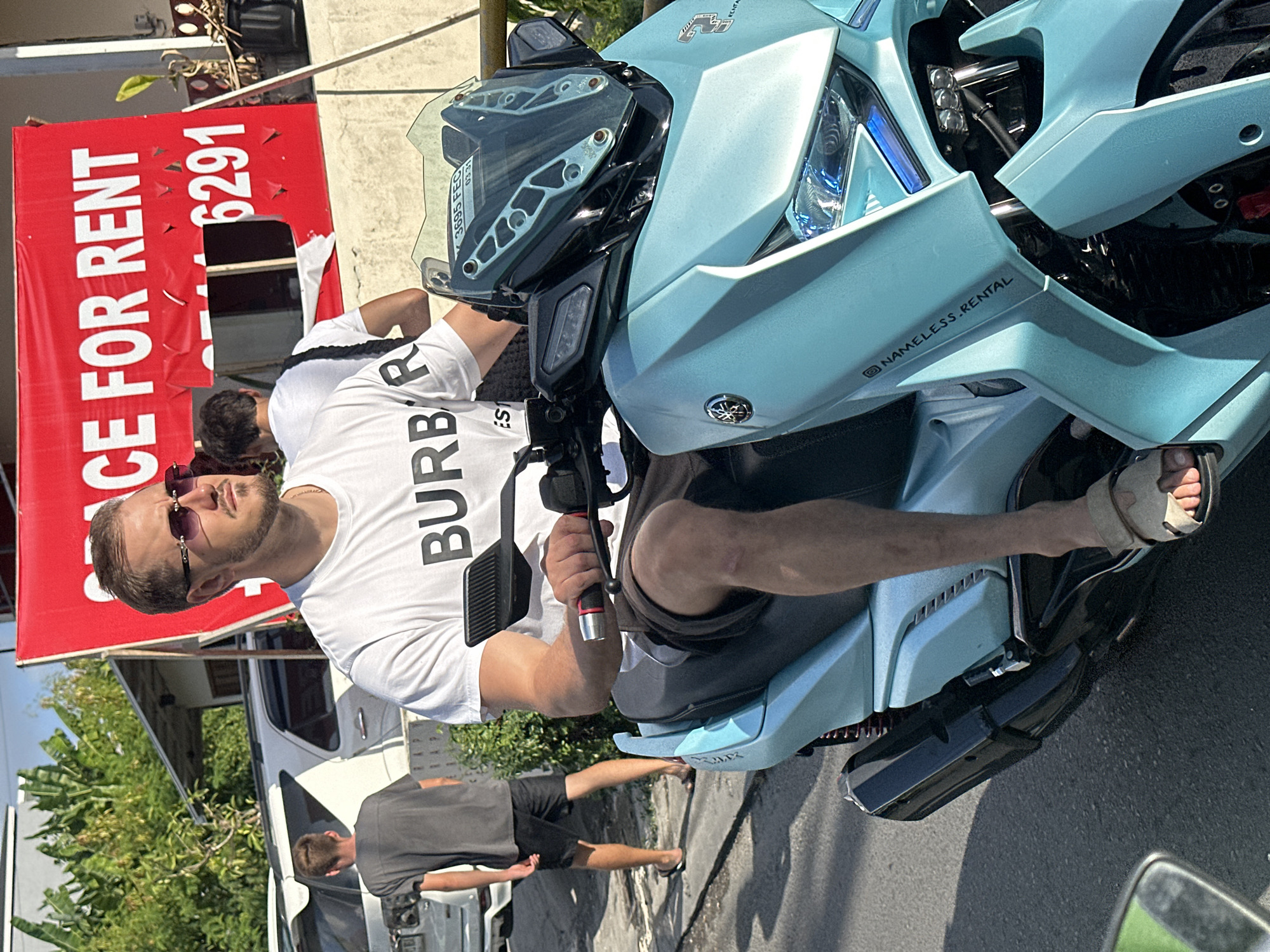 In North Kuta, Indonesia, a man with sunglasses and a white t-shirt with "BURBERRY" printed on it rides a light blue scooter. He is wearing dark shorts and sandals. To his left, another person in a dark t-shirt and shorts is walking away from the camera. Behind them, a white SUV is parked. A red banner with white text stating "CE FOR RENT" and a phone number is visible in the background, attached to a building with a concrete facade and a metal railing. The scene is illuminated by sunlight, casting shadows on the asphalt road.