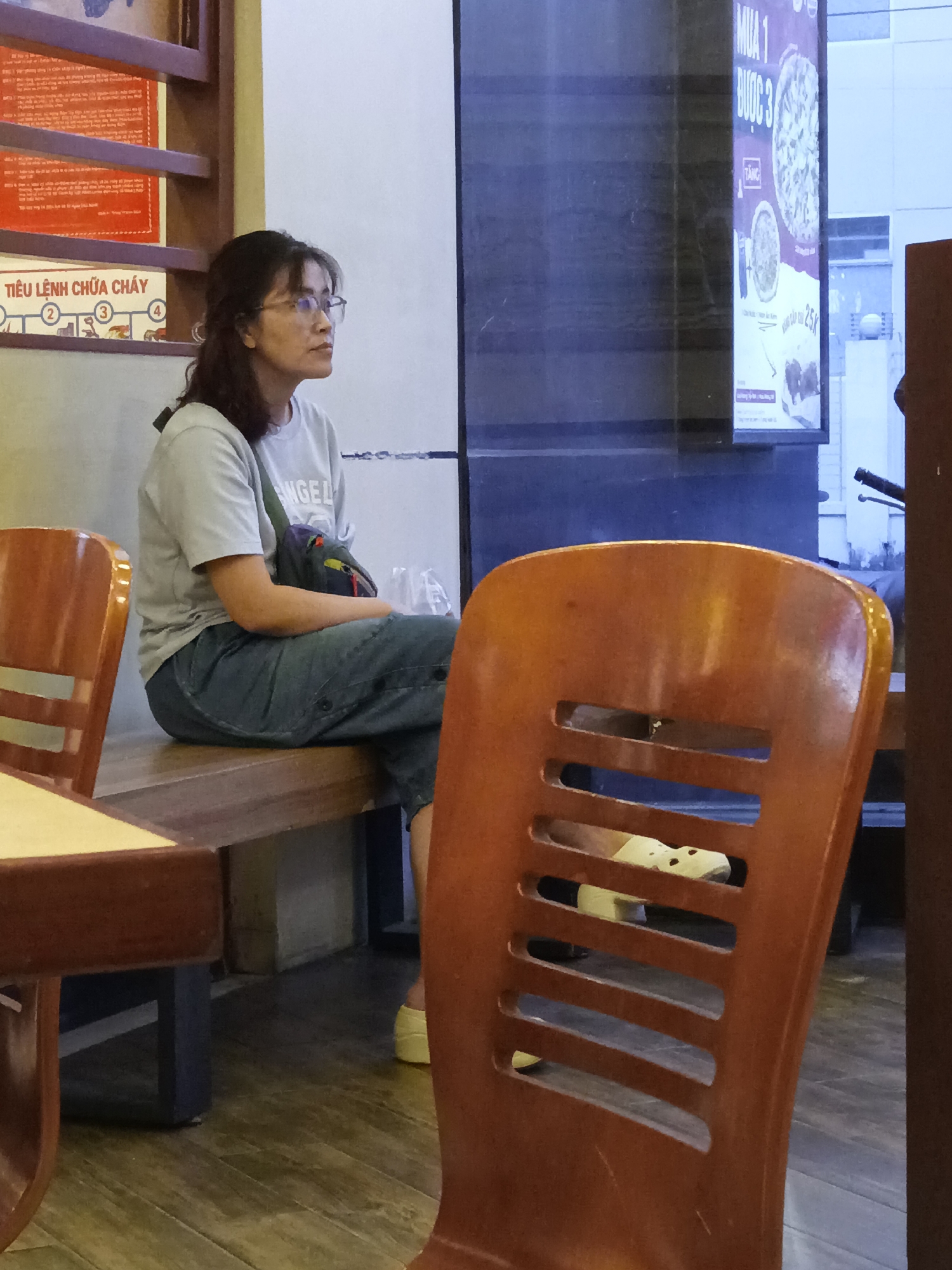 A woman is sitting on a wooden bench, looking to the right. She is wearing a light-colored t-shirt, blue jeans, and glasses. A bag is slung over her shoulder. In front of her, to her left, is a small table with a light-colored top and dark brown legs. To her right is a wooden chair with horizontal slats. In the background, a large poster with text and images is visible. The floor is made of wooden planks. This scene is located in Nha Trang Ward, Vietnam.