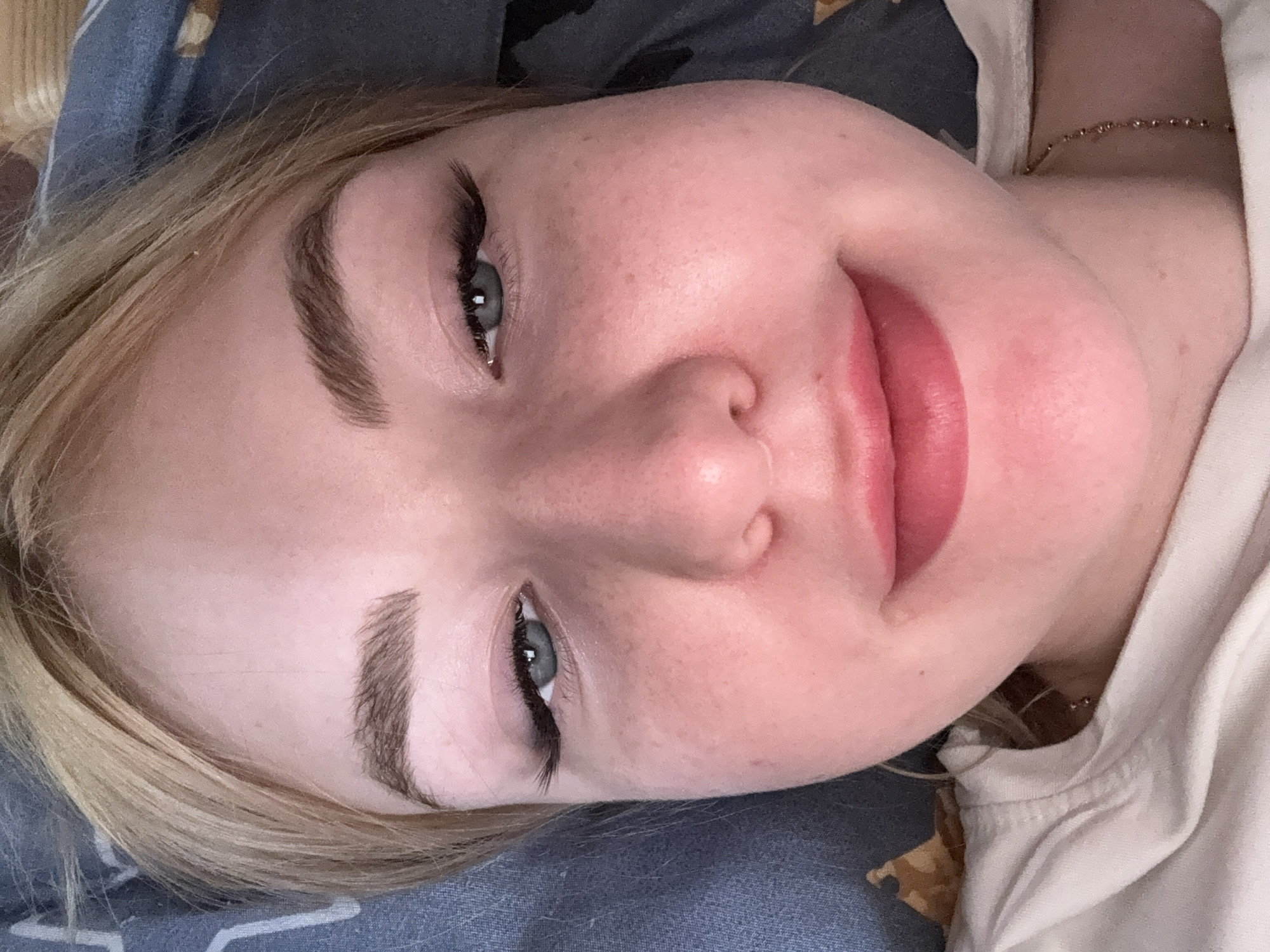 The image is a close-up, overhead shot of a young woman's face. She has fair skin with some freckles, blonde hair, and light blue eyes. Her eyebrows are well-defined, and she is wearing false eyelashes. Her lips are slightly parted and appear to be wearing pink lipstick. She is lying down on a blue and white patterned surface that appears to be bedding. There is a white t-shirt visible around her neck. The image is taken indoors, and the lighting suggests it is artificial. There is no discernible location in Krasnogorsk, Russia, as the image focuses solely on the subject's face and immediate surroundings.
