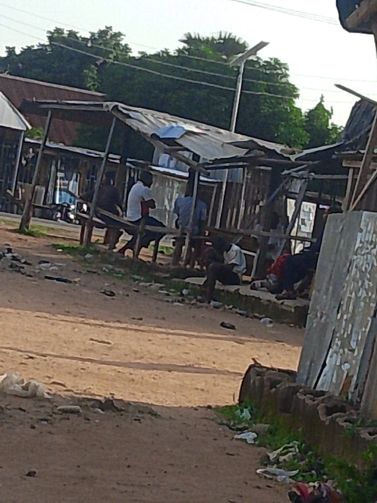 The image depicts an outdoor scene in Jalingo, Nigeria, likely during the daytime due to the bright sky and shadows. The setting appears to be a somewhat informal marketplace or gathering area with makeshift stalls constructed from wood and corrugated metal. Several people are present, mostly men, some sitting on wooden benches beneath the shelters and others standing or lying down.
In the foreground, a dirt road with some scattered debris suggests a rustic or possibly impoverished environment. Some white plastic bags and other refuse are visible on the ground, interspersed with sparse green vegetation.
The central focus is on the shelters where individuals are resting or socializing. One person in the mid-ground is wearing a white t-shirt and dark trousers, sitting on a bench. Others are also seated or lounging in various positions. The overall atmosphere seems relaxed, with no overt signs of urgency or distress, though the conditions of the shelters and the surrounding area might imply economic hardship.
In the background, trees and foliage indicate a natural environment surrounding the built structures. Power lines are also visible overhead, suggesting the presence of electricity. A vehicle, possibly a car, is partially visible on the road in the far left, adding a touch of everyday life to the scene. The lighting suggests it could be late afternoon or early morning, with the sun casting long shadows. There is no visible text in the image.