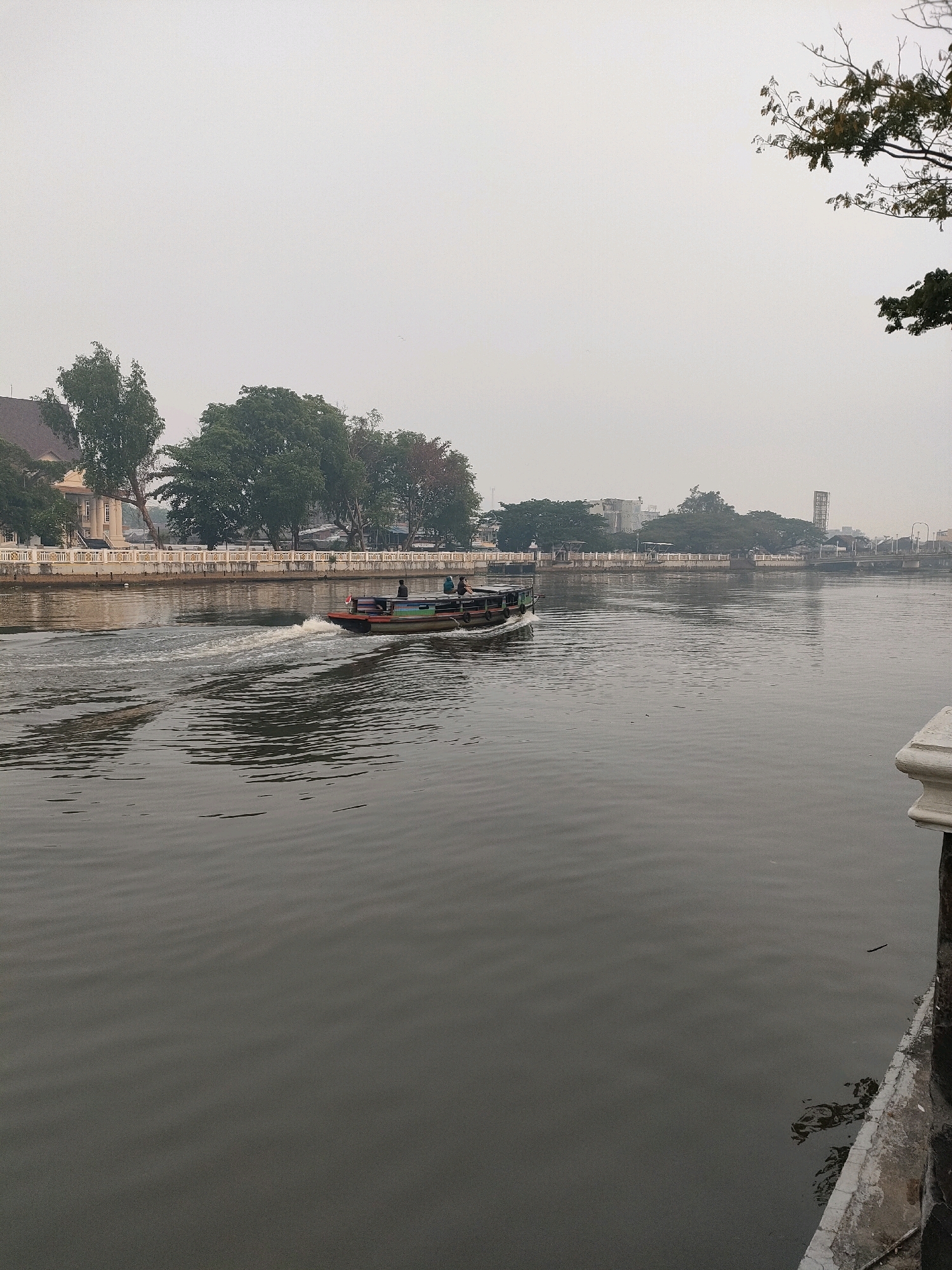 The image depicts a canal in Banjarmasin, Indonesia, characterized by a hazy sky and calm water. A long, narrow boat with a colorful stripe along its side is moving from left to right, leaving a wake in its path. Several individuals are visible on the boat. Along the left bank of the canal, there is a white railing bordering a walkway, beyond which are trees and buildings. The architecture of the buildings suggests a historical or colonial influence. The right side of the canal features a similar white railing and a concrete embankment. The overall atmosphere is muted, possibly due to overcast weather or mist.