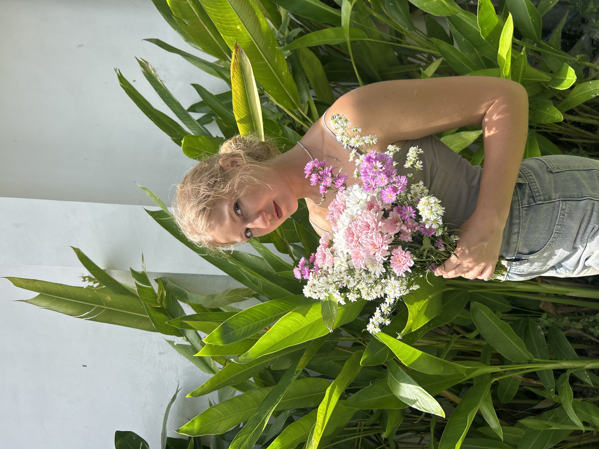 An outdoor, eye-level shot captures a blonde woman, likely in her 20s or 30s, holding a vibrant bouquet of flowers. Her hair is pulled back, revealing her fair skin and light-colored eyes, which look directly at the viewer with a calm expression. She wears a light-colored tank top and light blue denim jeans.
The bouquet, held in her hands, features a mix of pink, white, and light purple blooms, including what appear to be chrysanthemums and asters. Behind her, dense, lush green tropical foliage with large, elongated leaves creates a natural and vibrant backdrop. To her left, a plain white wall is visible.
The image is rotated 90 degrees clockwise from what would be a standard upright orientation, giving an unusual angle to the composition. Bright, natural sunlight illuminates the scene, highlighting the leaves and the woman's skin, suggesting a clear day in the morning or late afternoon. The setting, with its tropical plants, aligns with the location context of Kuta Selatan, Indonesia. There is no visible text in the image.