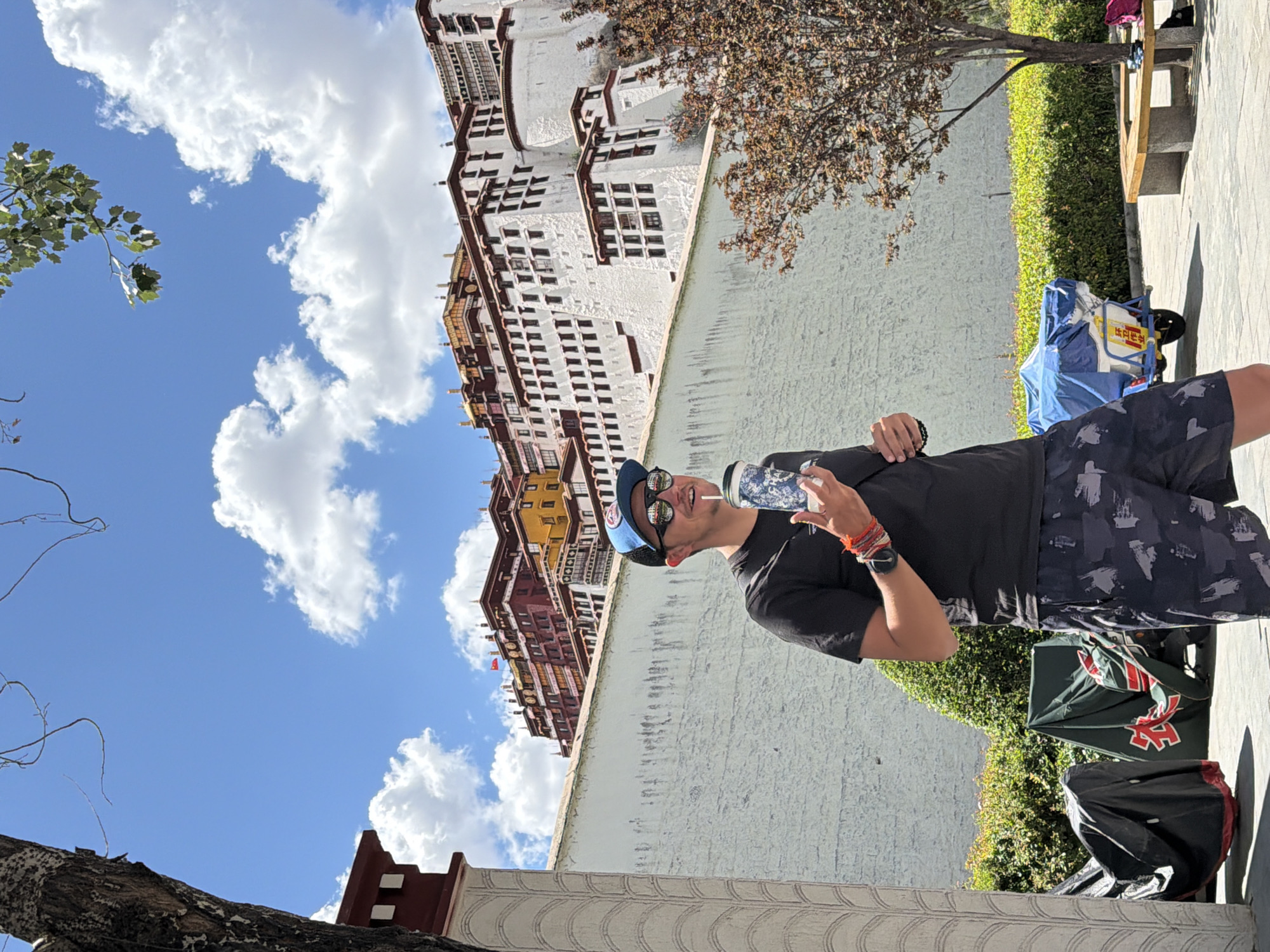 This outdoor image captures a bright, sunny daytime scene with a clear blue sky dotted with fluffy white clouds. The main subject is a man standing in the foreground, positioned slightly to the right of the frame. He is wearing a black t-shirt, patterned black and grey shorts, a blue baseball cap, and sunglasses, and he holds a patterned water bottle in his right hand. He has a slight smile on his face. Colorful bracelets and a watch are visible on his left wrist.
Behind the man, a light-colored wall runs horizontally across the mid-ground. Beyond the wall, a strip of vibrant green bushes and a tree with brown, autumn-like leaves are visible. To the left, a large, majestic building with distinct Tibetan architecture dominates the background. This multi-storied structure is predominantly white with red and dark brown sections, featuring many small, dark windows. Its appearance strongly suggests it is the Potala Palace in Lhasa, consistent with the Chengguan District, China location. Another tree with green foliage is visible in the upper left corner of the sky.
On the paved ground near the man's feet, to his right, are several items including a blue and white bag or piece of luggage, on which Chinese characters are visible, including "藏" (Zàng), a common character for Tibet. A black bag is also present nearby. Further to the right, a wooden bench is visible. The overall scene depicts a tourist enjoying a sunny day at a historic landmark.