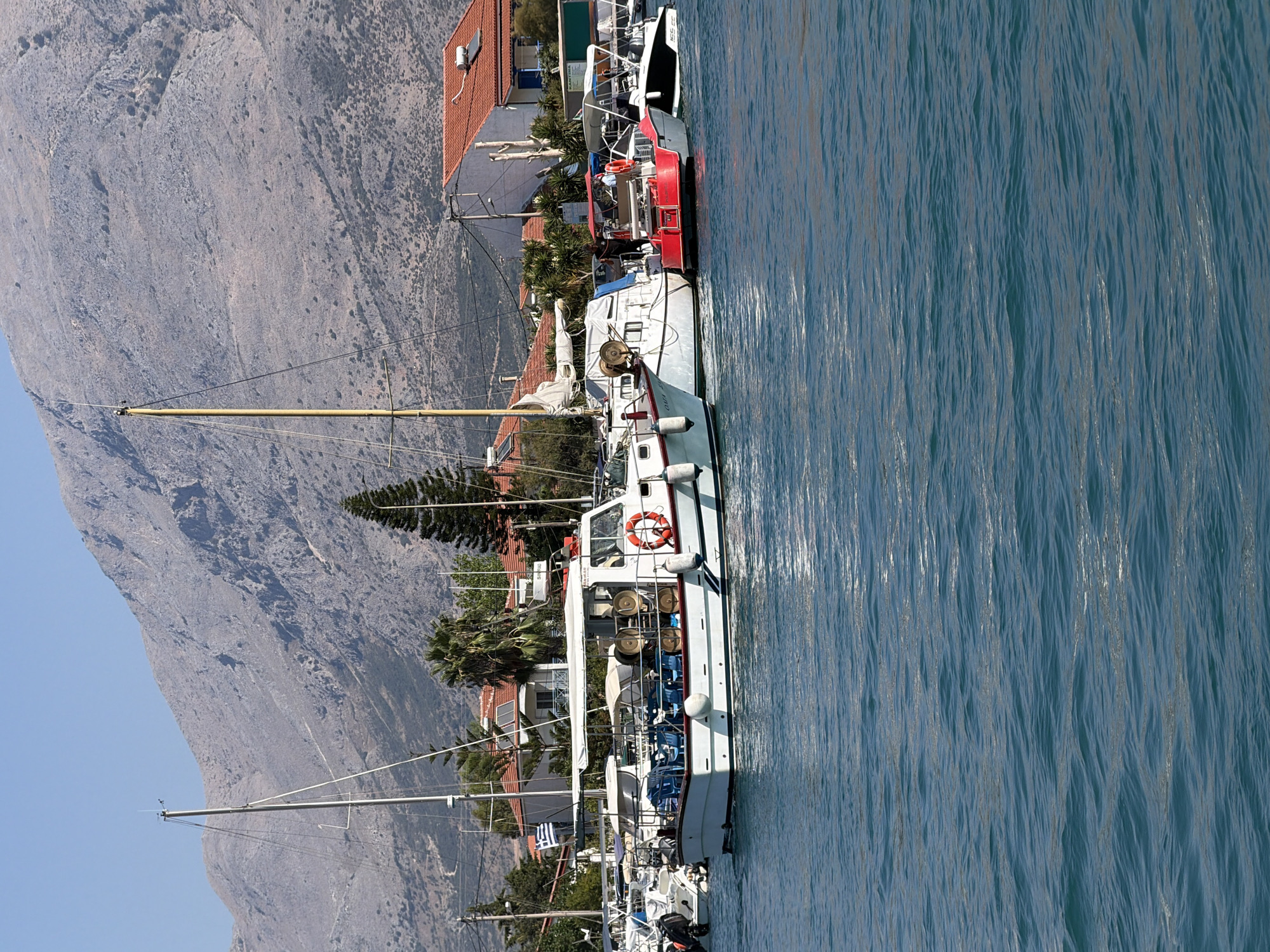 A fleet of moored vessels, including sailboats and motorboats, occupy the foreground of this image, set against the backdrop of the Municipal Unit of Alyzia, Greece. The water in the harbor is a vibrant blue, with gentle ripples reflecting the bright sunlight. Several boats are docked side-by-side, some with white canopies providing shade. One prominent motorboat, painted white with a blue stripe, has several chairs arranged on its deck and an orange life preserver attached to its railing. A red motorboat is moored next to it. In the middle ground, a cluster of buildings with red-tiled roofs are visible, interspersed with trees, including palm trees and a tall evergreen. A steep, rocky mountain dominates the background, its slopes sparsely covered with vegetation. The sky is a clear, bright blue. The scene depicts a tranquil harbor environment.