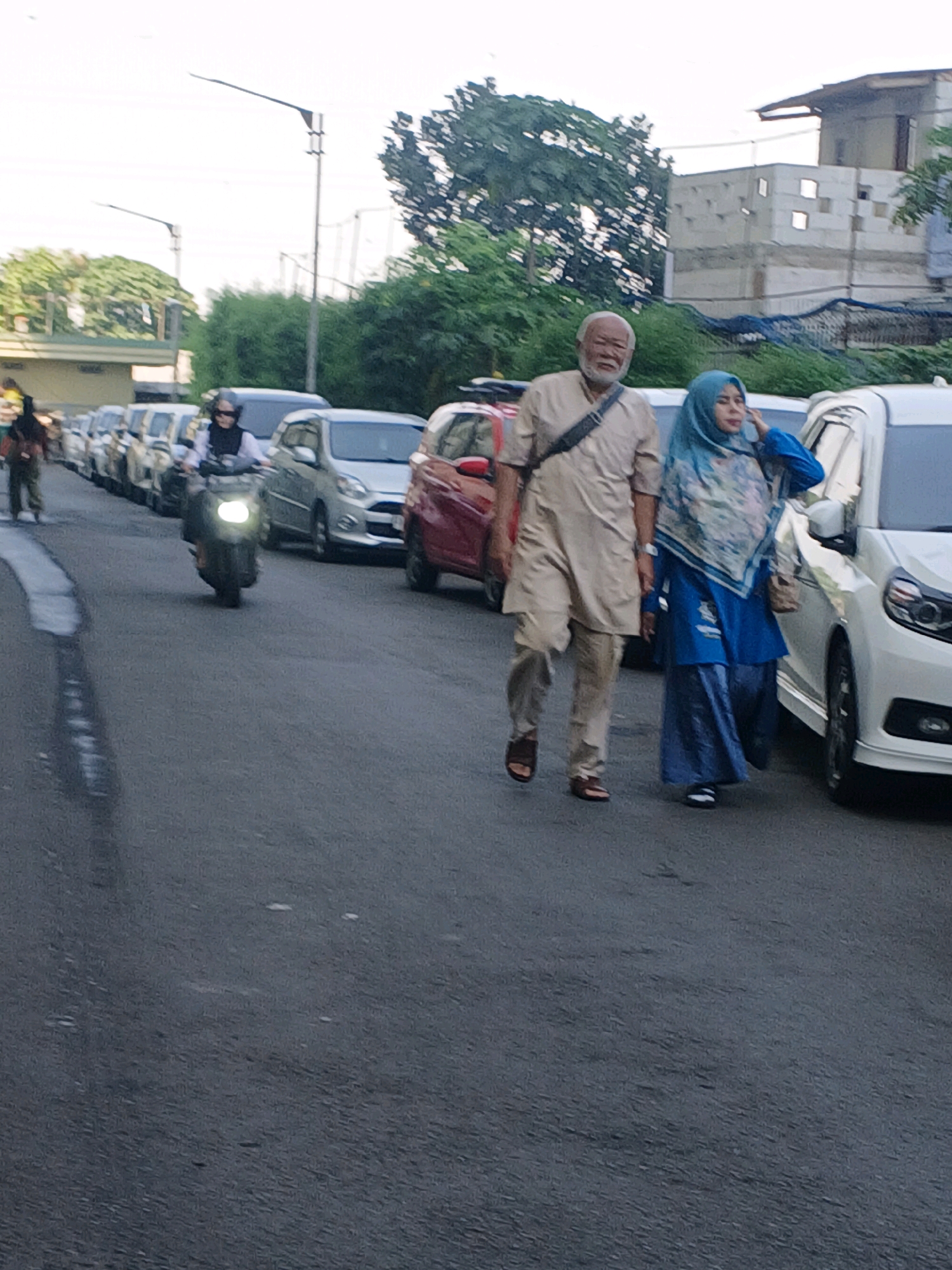 A low-angle, medium shot captures a street scene with several vehicles and pedestrians. In the foreground, an elderly man with a white beard, wearing a light tan tunic and pants, walks with a woman in a blue hijab and blue attire. They are walking alongside a white car. Further down the street, a person on a scooter with its headlight on is visible among a line of parked or slow-moving cars. In the background, trees and buildings, including a partially constructed building with visible scaffolding, are present. The road surface appears to be asphalt. The lighting suggests it is either early morning or late afternoon. The overall impression is a snapshot of urban life in Bandung City, Indonesia, with a focus on everyday activities and transportation.