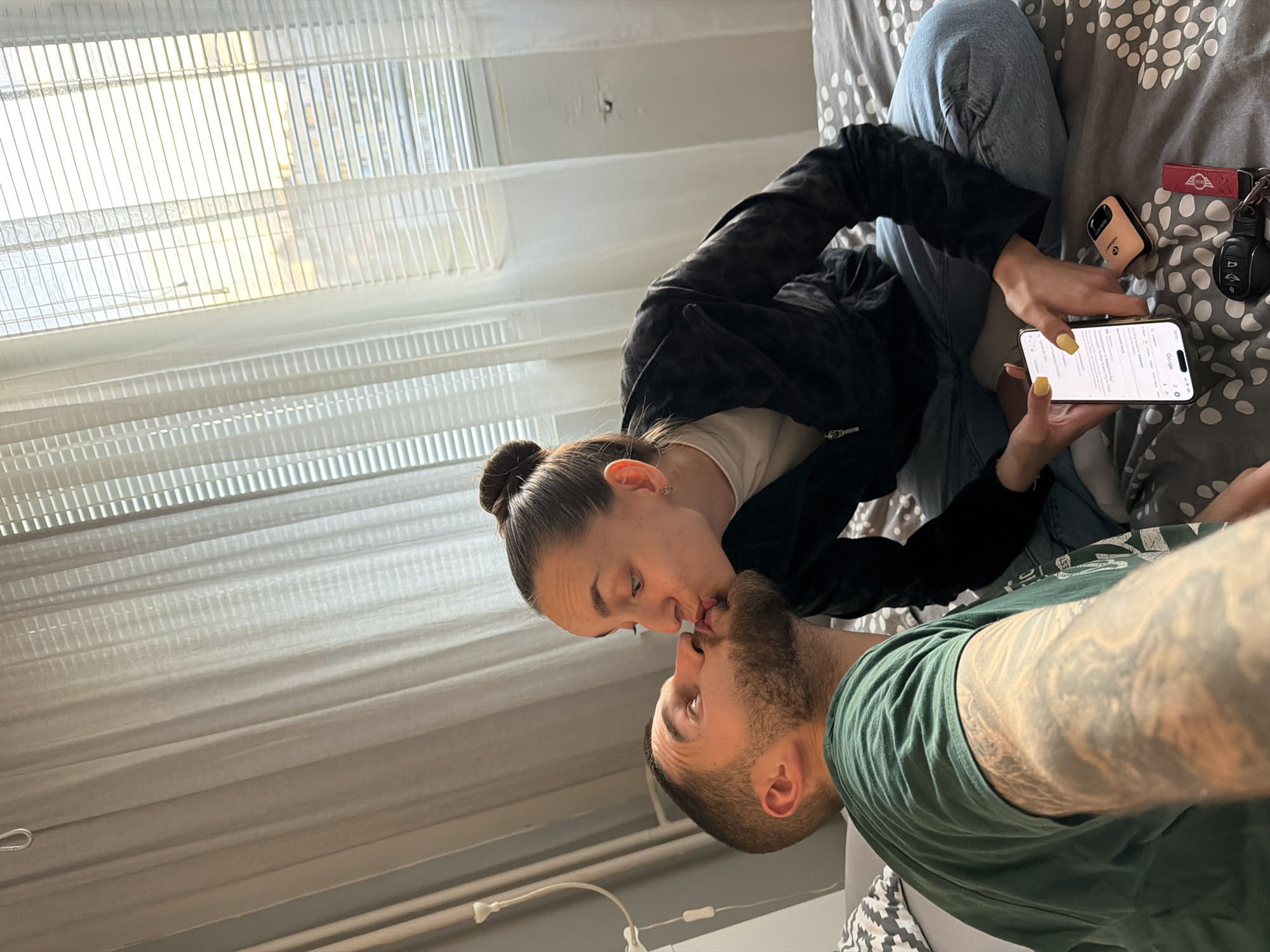 A couple is kissing while sitting on a bed. The woman has her hair in a bun and is wearing a black jacket and blue jeans, and she is holding a phone. The man is wearing a green t-shirt and has tattoos on his arm. The bed has a gray and white patterned blanket. A window with blinds is visible in the background. There is a lamp on the wall. The location is Sofia, Bulgaria.