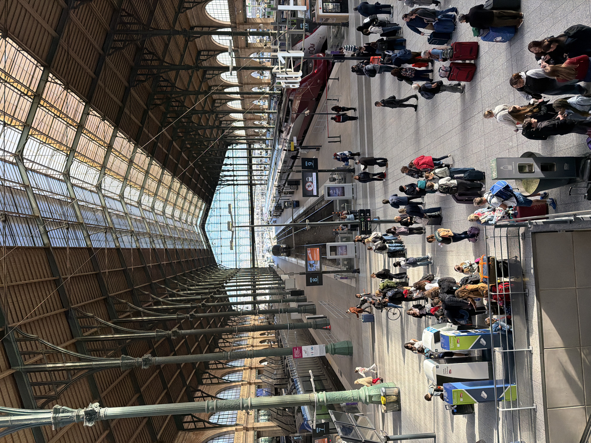 The image depicts the Gare de Lyon train station in Paris, France. The station's distinctive glass and iron roof structure is visible, with sunlight streaming through. Several train platforms are bustling with people, many of whom are pulling luggage. Some individuals are queuing near ticket barriers. A red train is parked at one of the platforms. Information boards and signage are present along the platforms.