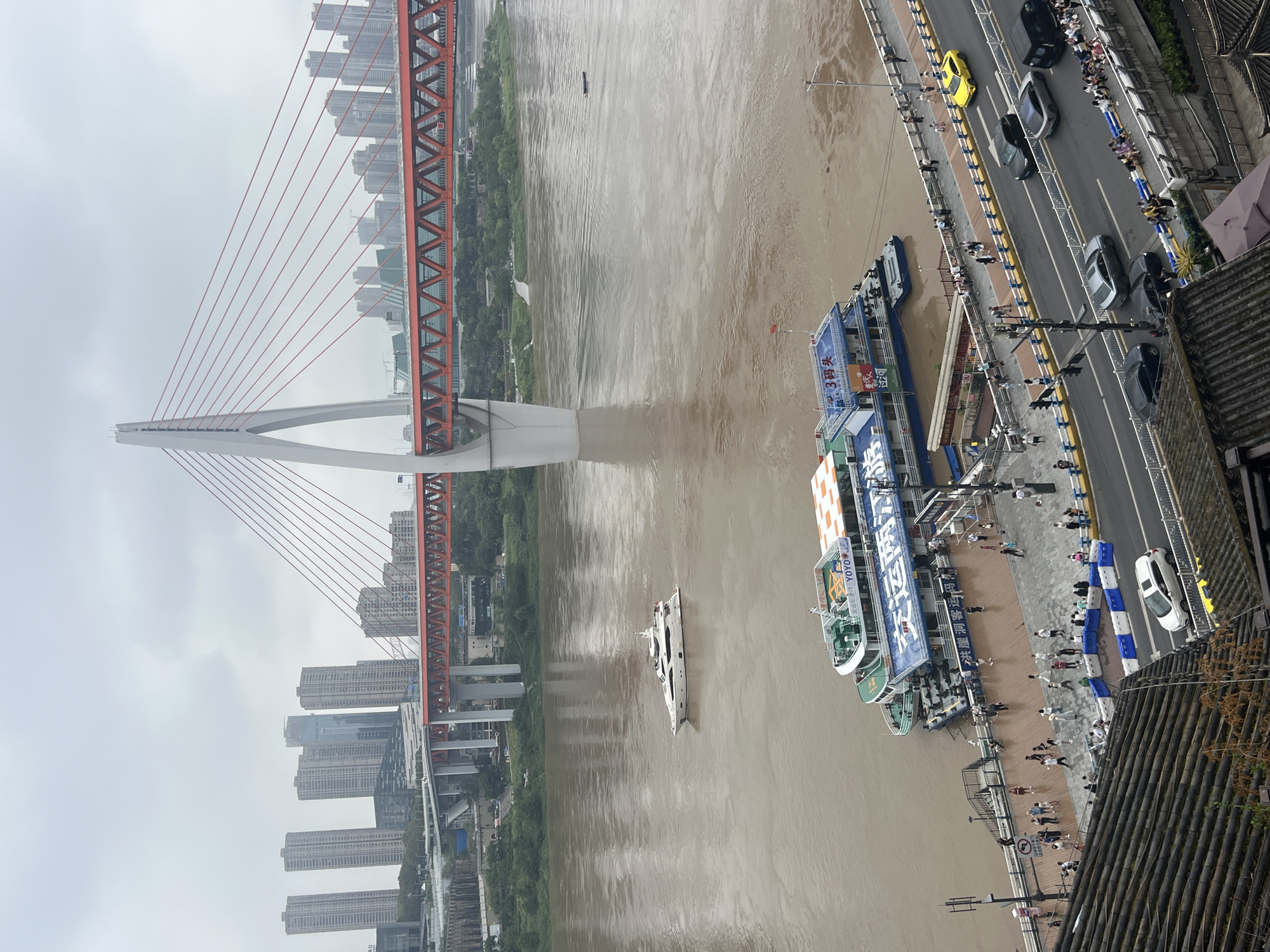 This image captures a view of the Yangtze River and the iconic Hongyadong Bridge in Chongqing, Yuzhong District, China. The sky is overcast, and the river is a murky brown color, indicating a high water level.
In the foreground, a traditional tiled roof is visible, suggesting a vantage point from an older building overlooking the modern cityscape.
Dominating the mid-ground are two passenger boats docked at a pier. The larger boat, painted in blue and white, displays the characters "3号码头" (Pier 3) and "游览观光" (Sightseeing). People are visible on the deck of the boats and along the wooden pier, some standing, others seated. A walkway lined with blue and white barriers separates the pier from a street above.
On the street, a yellow car is the most visible vehicle, traveling on the right side of the road. Several other cars, including black and silver sedans and a black van, are also present. A crowd of people are gathered along the railings of the elevated walkway beside the street, observing the scene below.
In the background, the Hongyadong Bridge spans the river. Its distinctive white pylon and red suspension cables are prominent against the cloudy sky. The bridge is a multi-level structure with red girders supporting the upper roadway. Beyond the bridge, a dense collection of modern high-rise buildings forms the city skyline. The overall impression is one of urban development juxtaposed with the powerful natural element of the river.