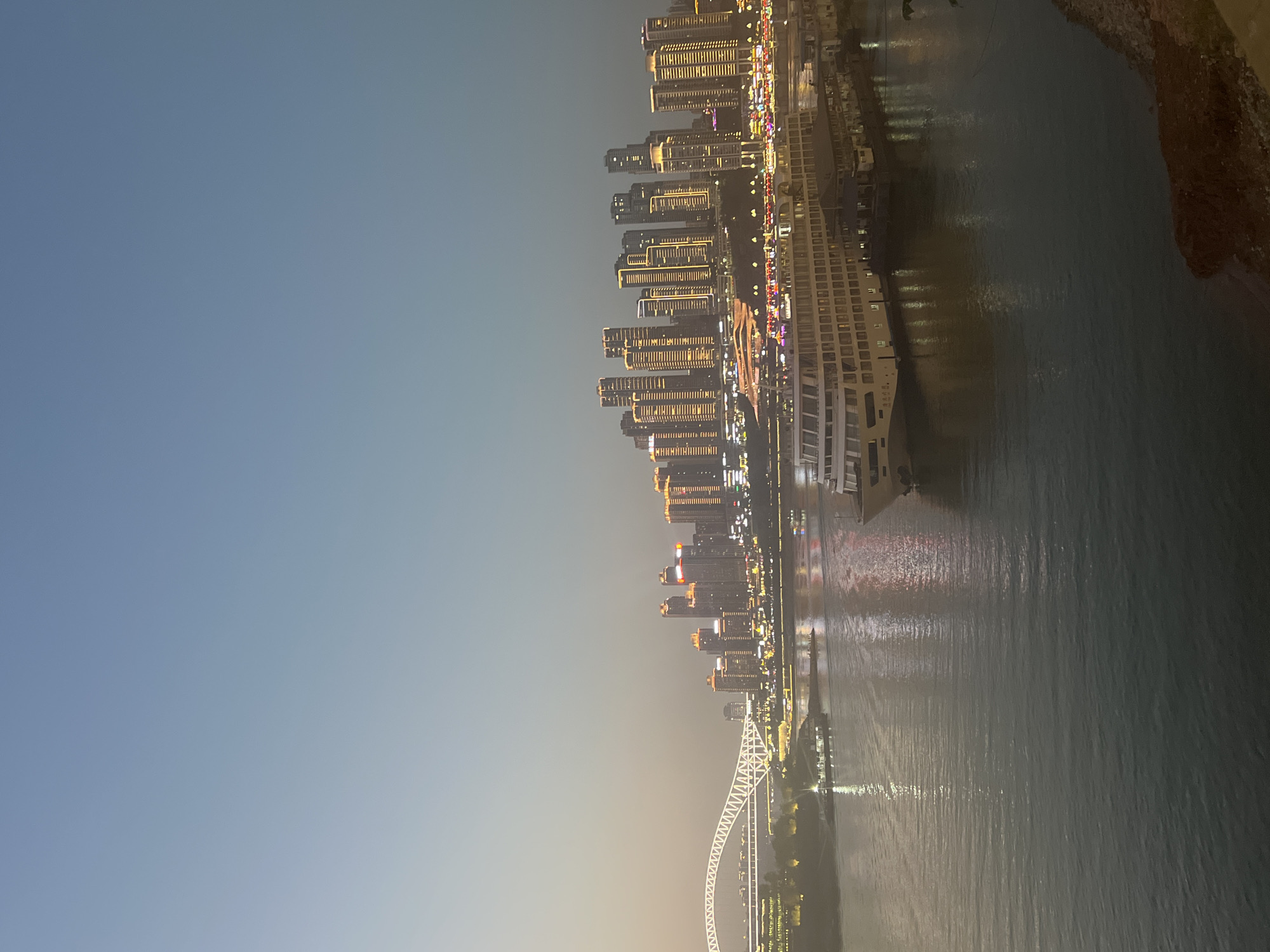 The image captures a wide-angle view of a city skyline at dusk, likely from Yuzhong District, China, given the prominent architecture. The sky is a soft gradient of pale blue to a hazy orange near the horizon, suggesting twilight. A large river dominates the foreground, its surface a muted gray-green with shimmering reflections of the city lights.
On the water, a large, multi-decked white cruise ship is docked on the right side of the frame. Its windows are illuminated with warm light, and its sleek design suggests modern passenger transport. Further to the left, smaller boats are visible on the river.
The city skyline is a striking feature, characterized by numerous tall buildings of varying heights and designs, all lit up with golden and orange lights, creating a vibrant contrast against the darkening sky. The architecture appears contemporary, with many structures featuring a stepped or tiered design.
A prominent white illuminated bridge with an arching structure spans the river on the left side of the image, connecting different parts of the city. Below the bridge and along the riverbank, more buildings and lights are visible, indicating a densely populated urban area. The overall scene conveys a sense of urban dynamism and the transition from day to night in a bustling city. There are no people clearly discernible in the image.