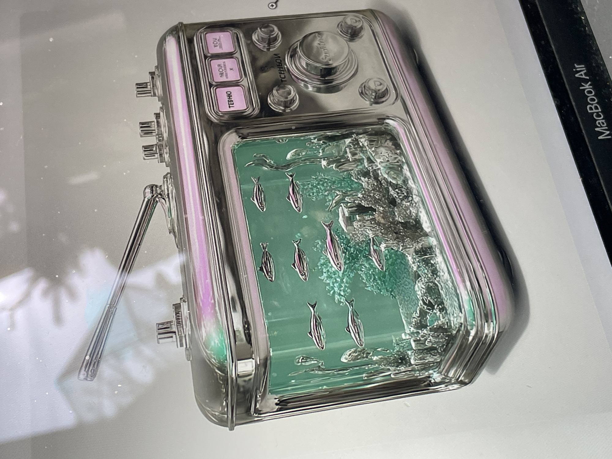 A close-up shot presents a silver, rectangular device with rounded edges, resembling a vintage radio or musical instrument. The device features a screen displaying an aquatic scene populated by several small fish. Above the screen, there are buttons and a knob labeled in a language that includes the Cyrillic alphabet, hinting at a Russian origin. The lighting casts a slight iridescence across the device's surface. The scene also includes a portion of a laptop with the words "MacBook Air" printed on its frame. It is possible this device is associated with the Sestroretsk region in Russia due to the Russian text on the device.