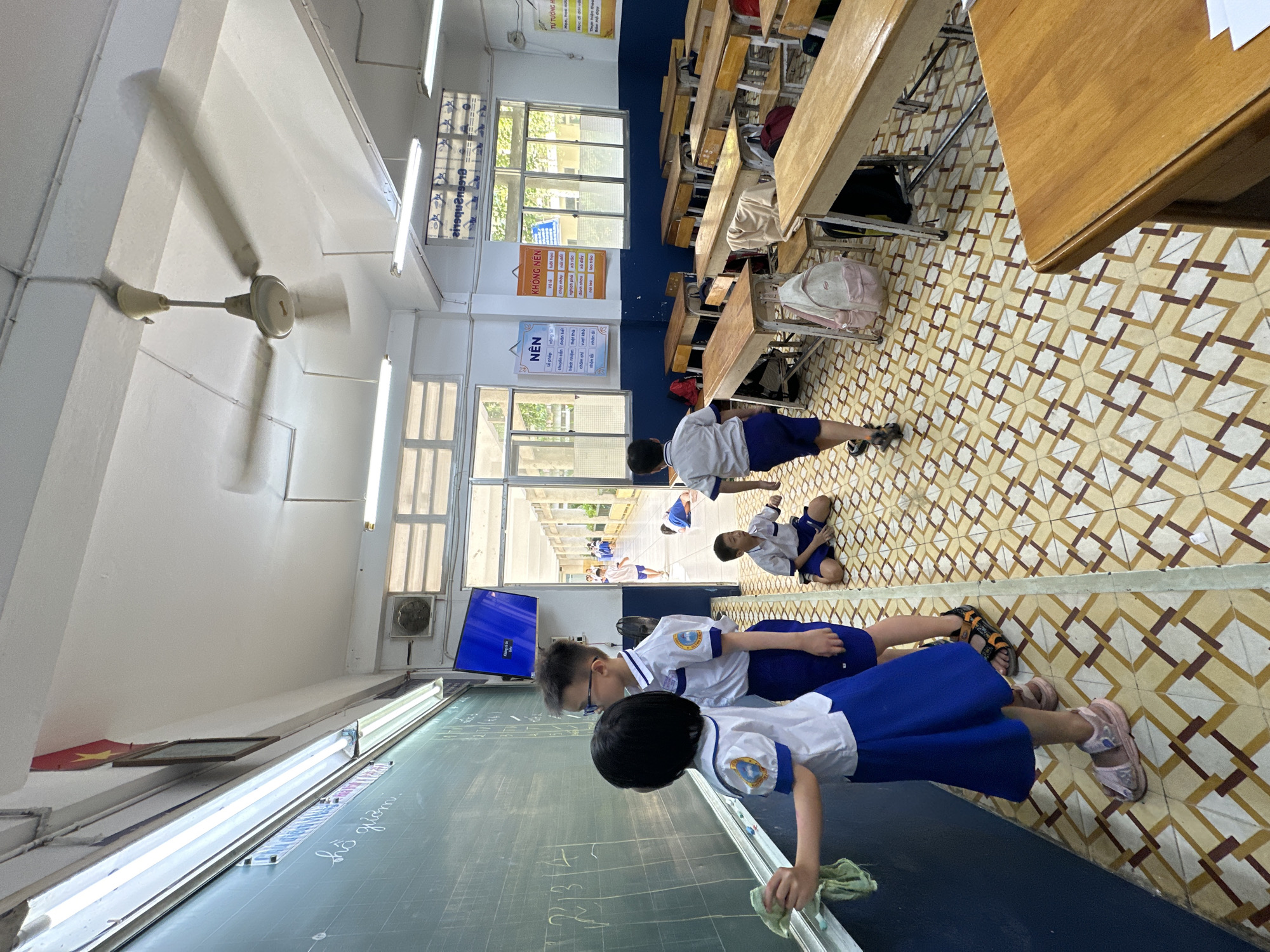 The image depicts the interior of a classroom in Ho Chi Minh City, Vietnam, during daytime. The room features a distinctive geometric tiled floor in shades of brown, yellow, and white, with a lower blue wall and white upper walls and ceiling. Fluorescent lights and a spinning ceiling fan illuminate the space.
Several students, dressed in white shirts with blue trim and blue bottoms (shorts or skirts), are present. In the foreground, a girl is actively wiping a large blackboard with a cloth, where chalk writing, including "hồ gươm." (Hoan Kiem Lake) and some numbers, is visible. A boy stands beside her, also facing the blackboard. Further back, other students are either standing near desks or sitting on the floor. Desks and chairs, mostly wooden, are arranged in rows, with some carrying school bags.
Bright natural light streams in through large windows on the left, revealing an outdoor school corridor or walkway with more buildings and a glimpse of greenery. A TV screen is mounted on the wall near the windows, currently displaying a blue screen.
Posters on the blue wall provide behavioral guidelines in Vietnamese. One poster under the heading "NÊN" (SHOULD) lists positive traits like "Lễ phép" (Respectful), "chuyên cần" (Diligent), "đoàn kết" (United), and "nhận lỗi" (Admit mistakes). Another poster under "KHÔNG NÊN" (SHOULD NOT) warns against actions such as "lười học" (Lazy in study), "chạy nhảy" (Run and jump), "nói dối" (Lie), "xả rác" (Litter), and "đánh nhau" (Fight). A small, partially visible Vietnamese flag is placed on a ledge above the blackboard. The overall atmosphere suggests a typical school environment, possibly during a cleanup activity or a break.