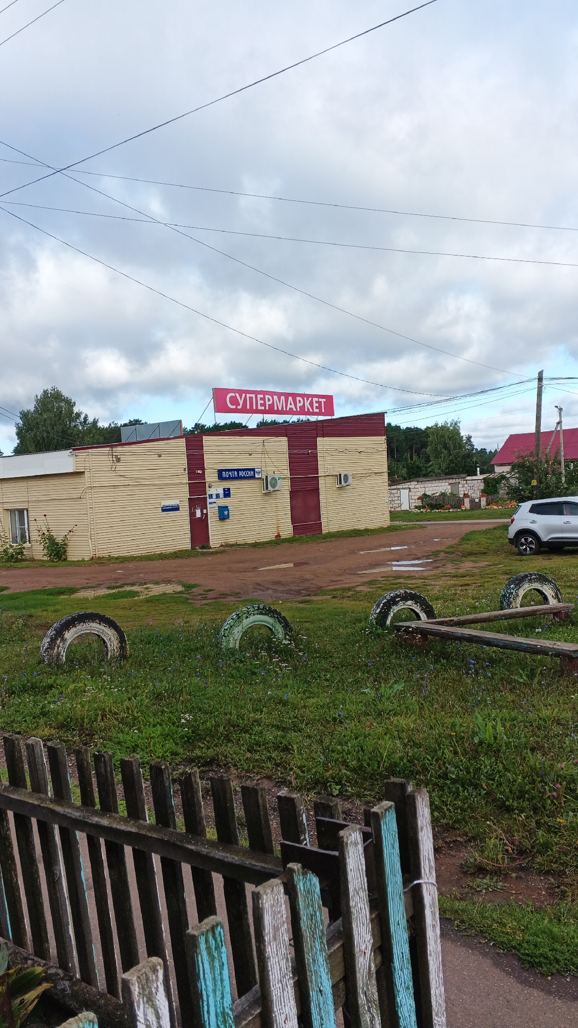 The image shows a low-rise building with a pink sign that reads "СУПЕРМАРКЕТ" (Supermarket) in Cyrillic letters. The building appears to be a commercial establishment, with a smaller sign below it indicating "ПОЧТА РОССИИ" (Russian Post). The building's facade is painted in alternating bands of yellow and dark red. There are two air conditioning units mounted on the exterior wall. In front of the building, there is a dirt path and an area of green grass.
In the foreground, there is a weathered wooden fence with vertical slats, some of which are painted with faded blue and white. Beyond the fence and in the grass, there are three old car tires, two of which are standing upright and one lying on its side. A simple wooden bench-like structure made of two planks resting on what appear to be tire supports is also present in the grassy area.
A silver car is parked on a paved area to the right of the building. In the background, there are trees and other buildings with red roofs, suggesting a rural or semi-rural setting. Overhead, a network of power lines is visible against a cloudy sky. The overall scene depicts a common commercial and postal facility in a village or small town, likely in Russia given the Cyrillic signage. The location appears to be the village of Podgornoye, Russia, based on the provided context.