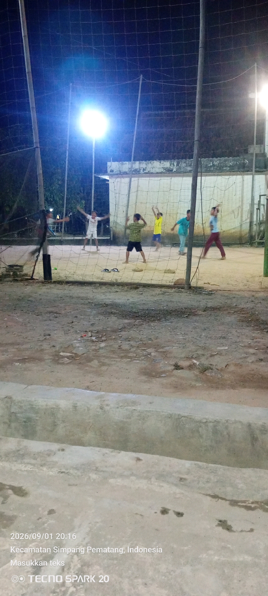 The image depicts an outdoor volleyball court at night, illuminated by bright floodlights. Several individuals, appearing to be young males, are on the court, some actively engaged in playing volleyball and others in various athletic poses. The court surface appears to be sand or dirt, and a net divides the playing area. In the foreground, a concrete barrier is visible, and beyond that, the ground is composed of dirt and scattered debris. The background shows a low building structure and trees. The lower portion of the image contains text indicating the date and time as "2026/09/01 20:16", the location as "Kecamatan Simpang Pematang, Indonesia", and a brand logo "TECNO SPARK 20".