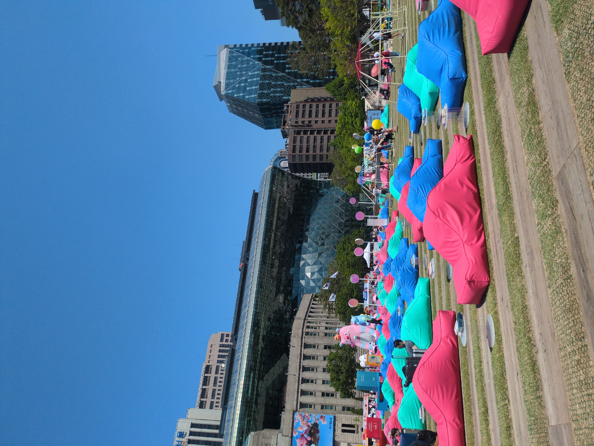 The image shows an outdoor event in Seoul, South Korea. Rows of brightly colored beanbag chairs, primarily pink, blue, and green, line a grassy area interspersed with wooden planks. People are sitting on or near some of the chairs. The urban setting includes modern skyscrapers in the background. Flags, balloons, and decorative elements such as cut-out figures add to the event's atmosphere.
