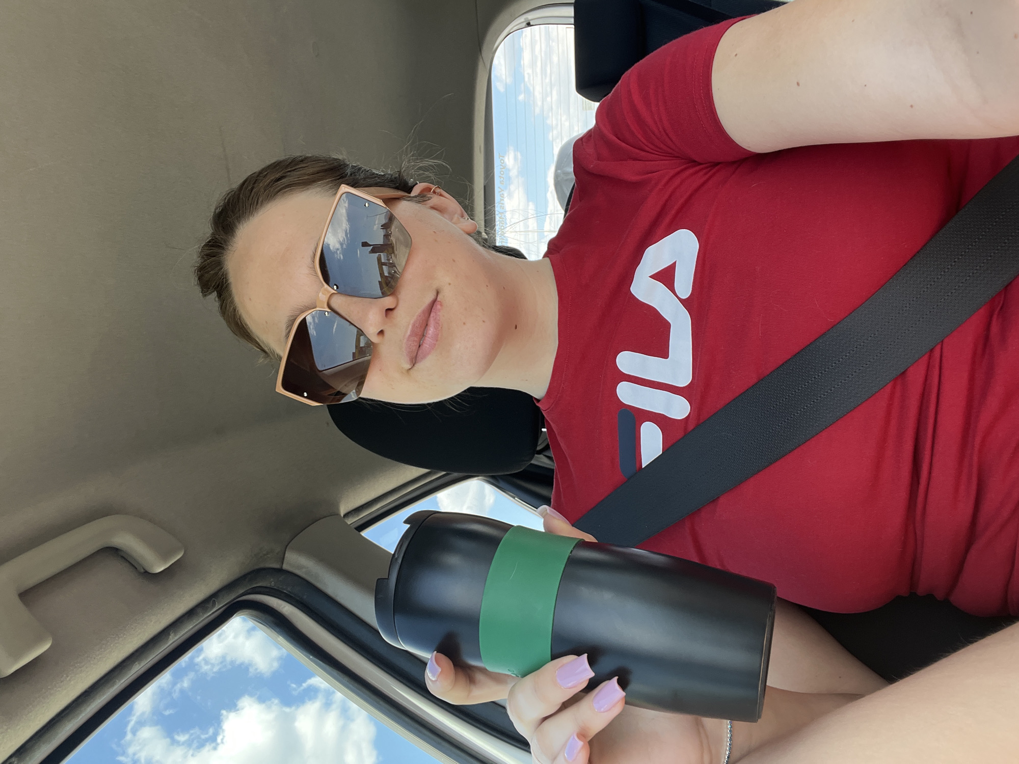 A woman wearing a red Fila t-shirt and sunglasses is seated in a car, wearing a seatbelt, and holding a black travel mug with a green band around it. The view through the car window shows a blue sky with white clouds. The photo appears to be taken inside a car, not in Donja Cijevna, Montenegro.
