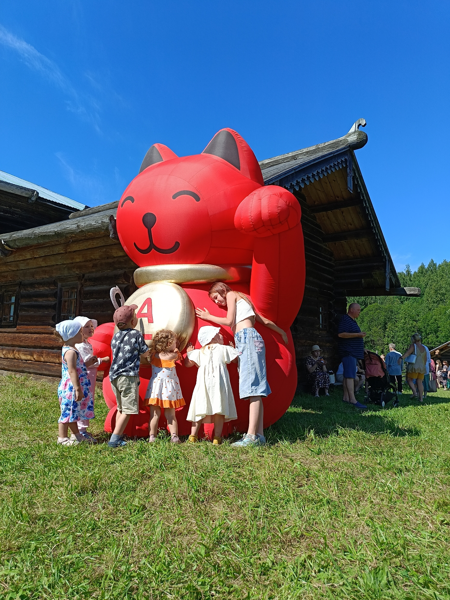 The image captures a sunny outdoor scene, possibly at a festival or event, featuring a giant inflatable red cat with its arm raised. A group of five young children, dressed in casual summer attire, are gathered around the base of the inflatable, some touching it and looking up at it. A young woman is standing behind the children, also interacting with the inflatable. In the background, a rustic wooden building with a distinctive roofline stands prominently. Several other adults and a stroller are visible further in the background, along with lush green grass and trees. The sky is a clear, vibrant blue with a few wispy clouds.