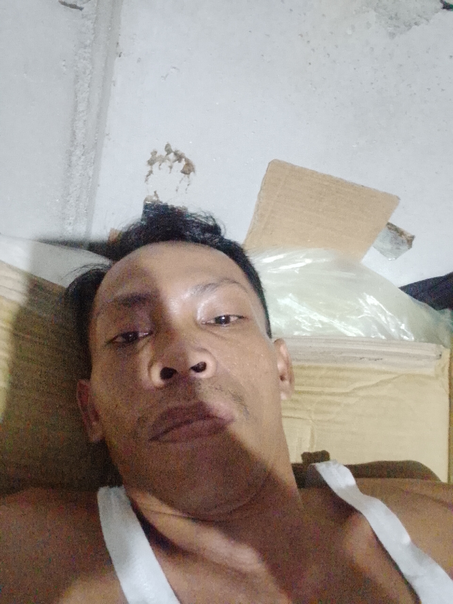 A man is lying down, facing upwards towards the camera. He is wearing a white tank top. Above him are pieces of cardboard and a clear plastic bag. The location is a room with a white wall and ceiling. Bukit Ketok, Indonesia, is specified as the location.