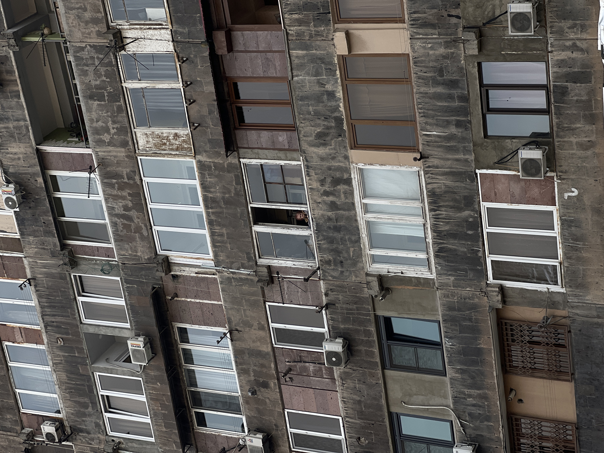 A full shot of an old multistory building with numerous windows. The building is made of a dark, possibly stone material, and appears to be weathered. The windows are of varying styles and ages, some with air conditioning units attached. In one of the windows, a person is visible. The location appears to be an urban area, though the specific city is unknown.