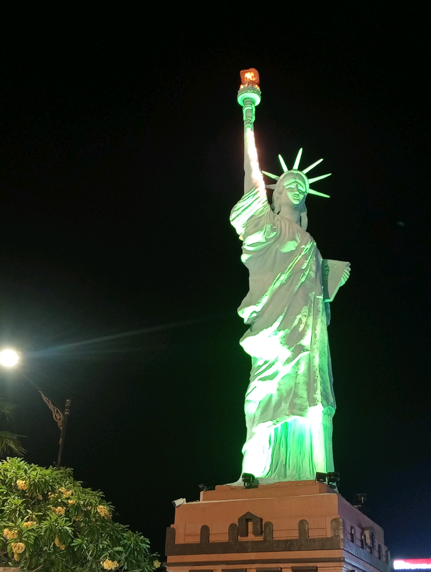 The image shows a replica of the Statue of Liberty illuminated by green lights at night. The statue is standing on a pedestal with ornate architectural details. To the left of the statue, there is a bright light source that appears to be a streetlamp, and a tree with yellow flowers. In the background, there is a dark sky with some faint lights. This replica of the Statue of Liberty is located in Madiun, Indonesia, a tourist attraction known as the "Patung Liberty Madiun".
