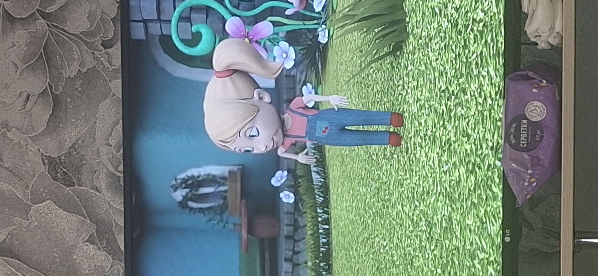 The image shows a 3D animated cartoon character of a young girl with blonde hair tied in a ponytail, wearing a pink shirt and blue overalls with cherry designs on the pocket. She is standing in a grassy area with several small blue and purple flowers. In the background, there is a blue wall, some stone steps, and decorative green swirling structures with purple flowers. To the left of the character, there is a red round table and a green bench. The bottom of the image shows a purple package with Russian text that appears to be wet wipes. The LG logo is visible on the left side of the bottom edge. The scene is indoors, possibly on a screen, as indicated by the presence of a television screen frame and a patterned wallpaper above it. There is no indication of this scene being located in Bila Tserkva, Ukraine.