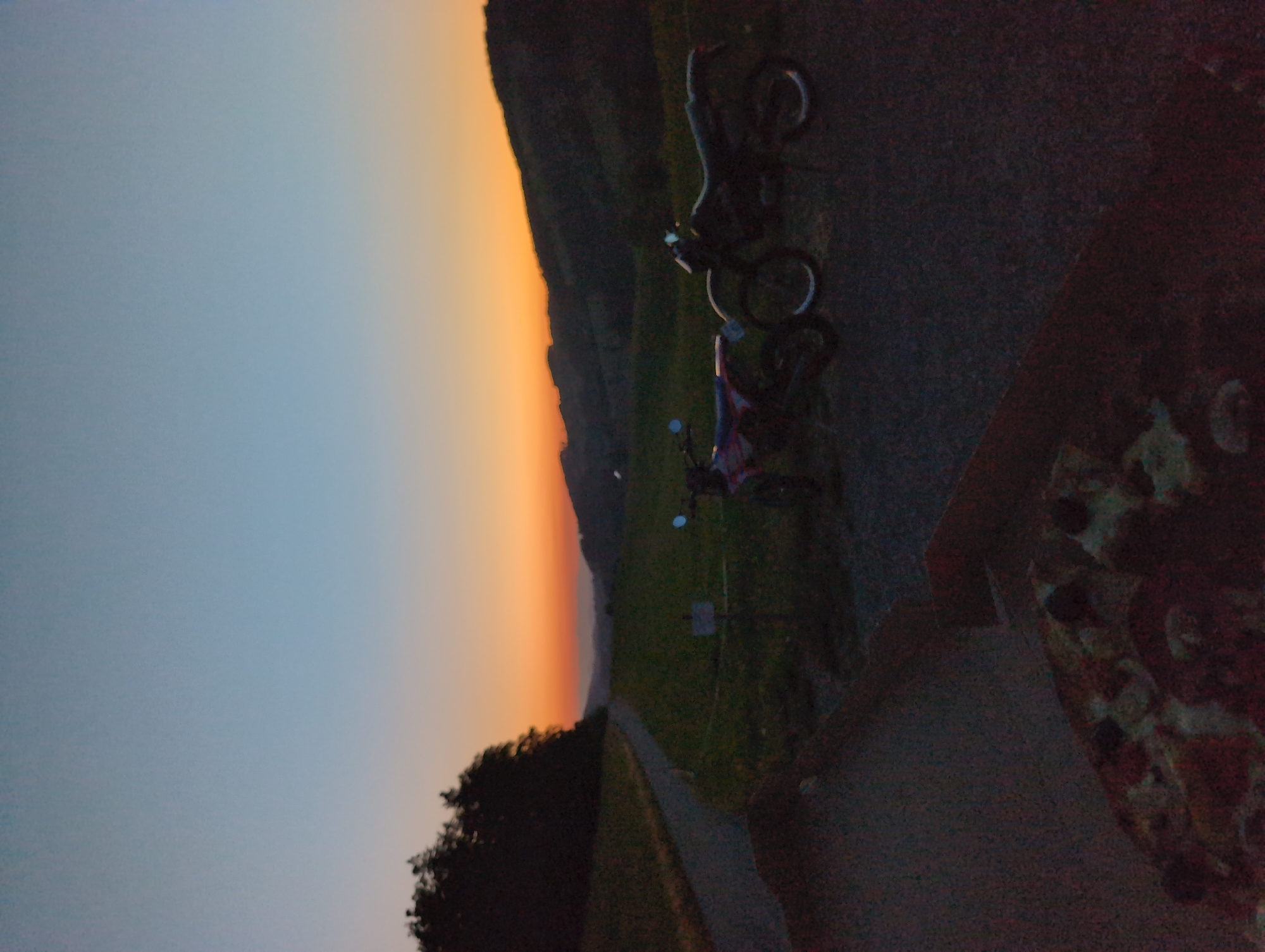 At dusk, two dirt bikes are parked on a gravel road in a rural area of St. Peter, Germany. The sky displays a gradient of blue to orange hues as the sun sets behind rolling hills covered in trees and greenery. In the foreground, a partially visible pizza box rests on the road. A paved path winds through a grassy field to the left of the motorcycles, bordered by trees. The lighting suggests late evening, with a clear sky.