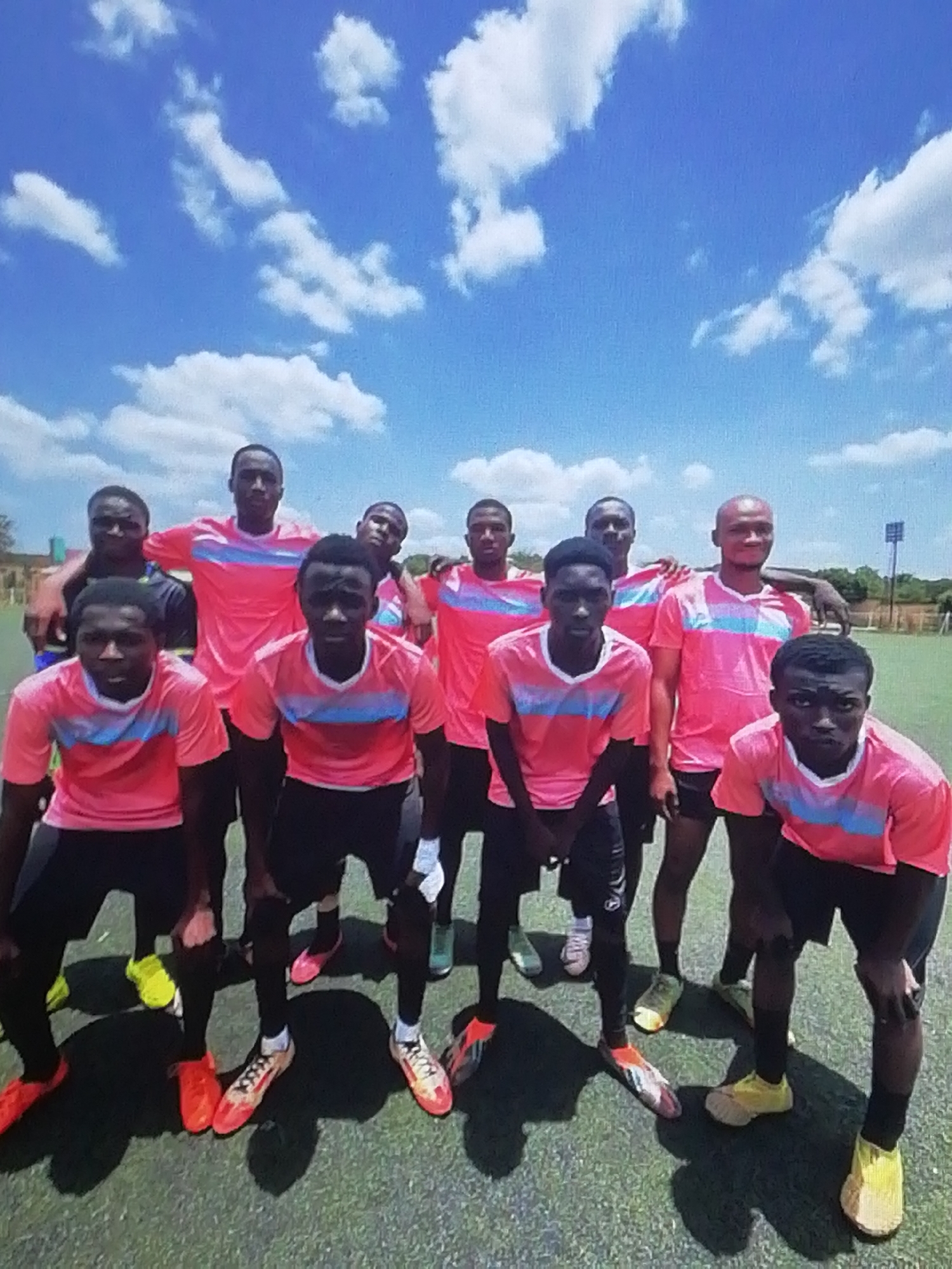A group of 10 young men pose for a team picture on a grass sports field under a bright, cloudy sky. They wear matching pink and light blue striped shirts with black shorts and a variety of colorful athletic shoes. The background features green trees and a structure resembling a light pole. While the image location is unknown, if this is the city of Enugu, Nigeria, it is not discernible from this image.