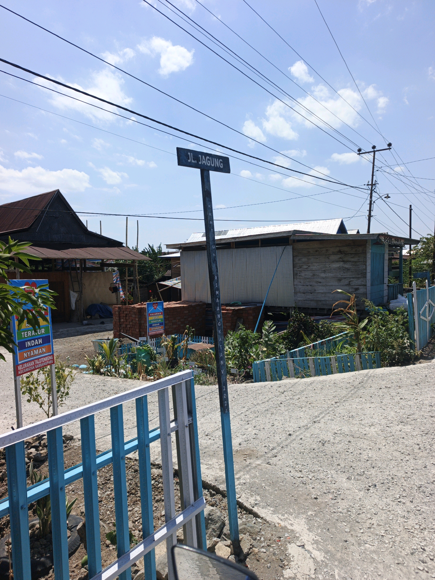 The image shows a street sign in Talotenreng, Indonesia, indicating "JL. JAGUNG". The sign is a dark rectangle with white lettering, mounted on a dark pole. The street is paved with gravel and appears to be in a somewhat rural or underdeveloped area, with several simple buildings visible in the background. One building has a dark, corrugated metal roof, and another is made of wood. There are piles of bricks and construction materials present. A blue and white fence runs along the side of the road. The sky is blue with scattered white clouds, and there are numerous power lines crisscrossing the scene. A small sign with colorful text and a heart symbol is also visible near the left side of the image, reading "TERA.UR INDAH NYAMAN KELURAHAN TALOTENRENG KECAMATAN SABBANPARU". There are no people visible in the image. The overall impression is of a sunny day in a developing community.