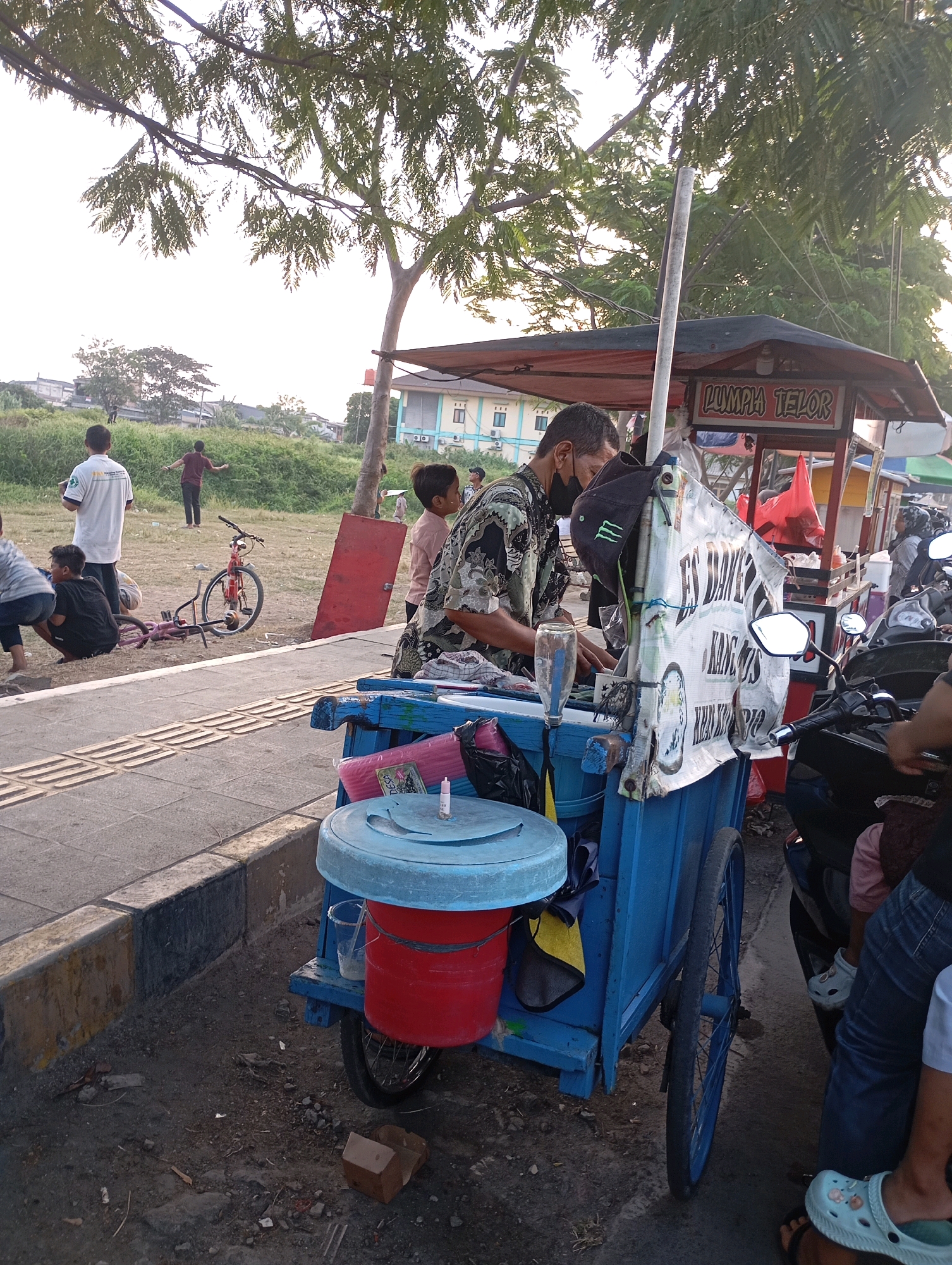 The image depicts a street scene in Babelan, Indonesia, with a focus on a food cart vendor. The vendor, a man wearing a patterned shirt and a black face mask, is standing behind a blue cart. The cart is equipped with a large blue lid on top, a red bucket below it, and a clear plastic cup containing a light-colored liquid. A sign on the cart reads "LUMPIA TELOR." A black baseball cap with a green logo is hanging from the cart. Several motorcycles are parked around the cart, suggesting a busy area. In the background, children and adults are visible, some walking and others sitting. A red bicycle lies on its side in the grass. The environment appears to be a park or recreational area, with trees and green spaces visible. The ground is a mix of paved walkways and dirt. The lighting suggests it is late afternoon or early evening.