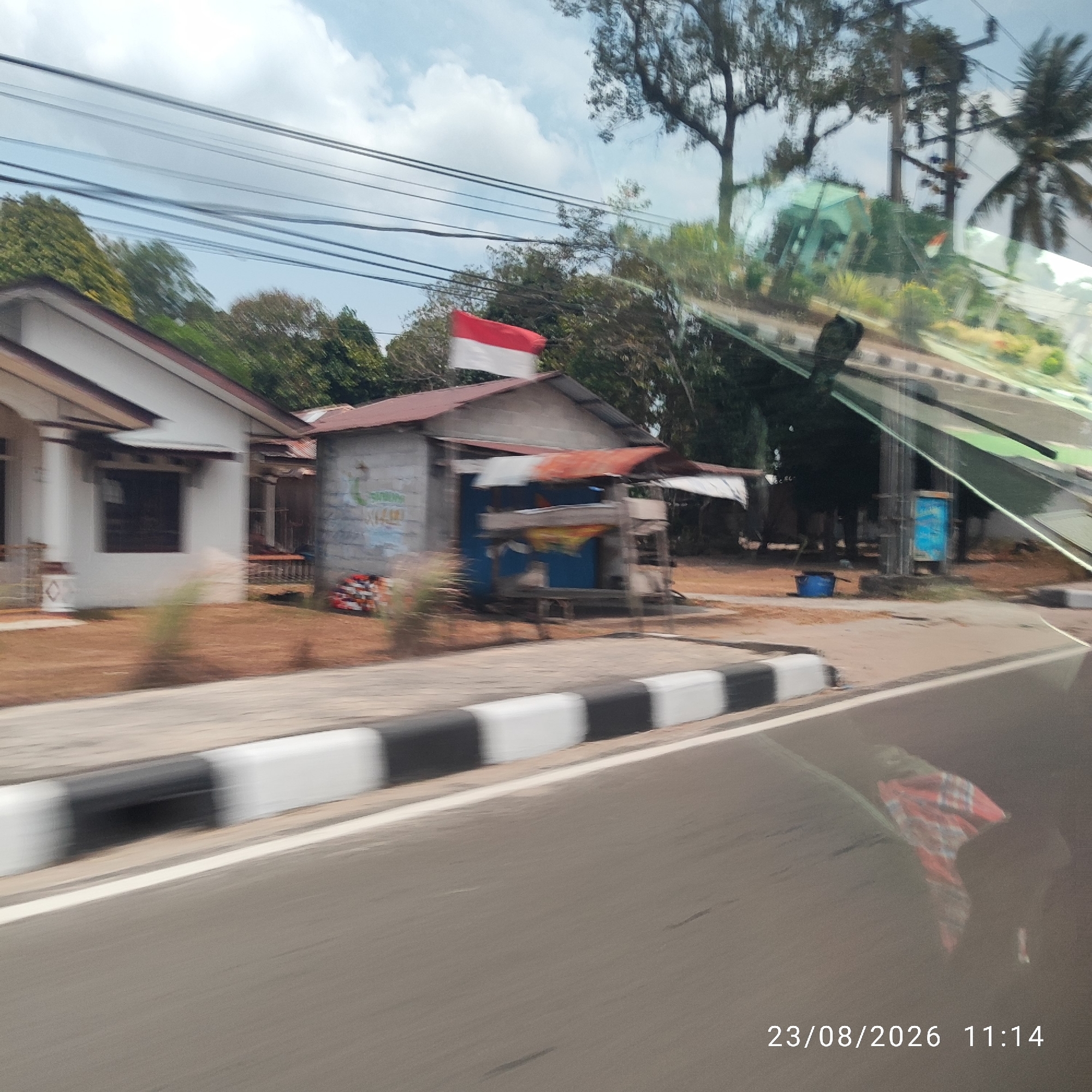 The image depicts a street scene in Tanjung Pandan, Indonesia. A red and white Indonesian flag is prominently displayed on the roof of a small, rustic structure, possibly a shop or a small dwelling. The structure itself appears to be made of corrugated metal and cinder blocks, with a simple wooden stall in front. Adjacent to this structure is a more modern, white house with a tiled roof. The street in front of these buildings is paved and has a distinct black and white striped curb. Overhead, several power lines crisscross the sky. The environment suggests a tropical or subtropical climate, with lush green trees visible in the background. The overall impression is one of a developing urban or semi-urban area. A timestamp in the lower right corner indicates the image was taken on 23/08/2026 at 11:14.