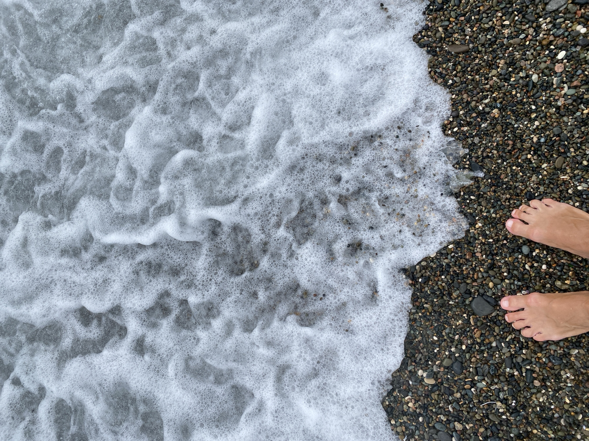 A pair of bare feet stands on a pebble beach as a wave with white foam recedes. The feet are positioned at the bottom of the frame, with the toes of the left foot slightly angled inward and the toes of the right foot angled slightly outward. The pebbles on the beach are of various sizes and colors, ranging from dark gray to light brown and white. The water is a light gray color and is filled with small bubbles. The scene appears to be a close-up shot, with the focus on the feet and the water's edge. The overall impression is one of a tranquil and serene moment at the seaside. The location is the shore of Sokhumi, Abkhazia.