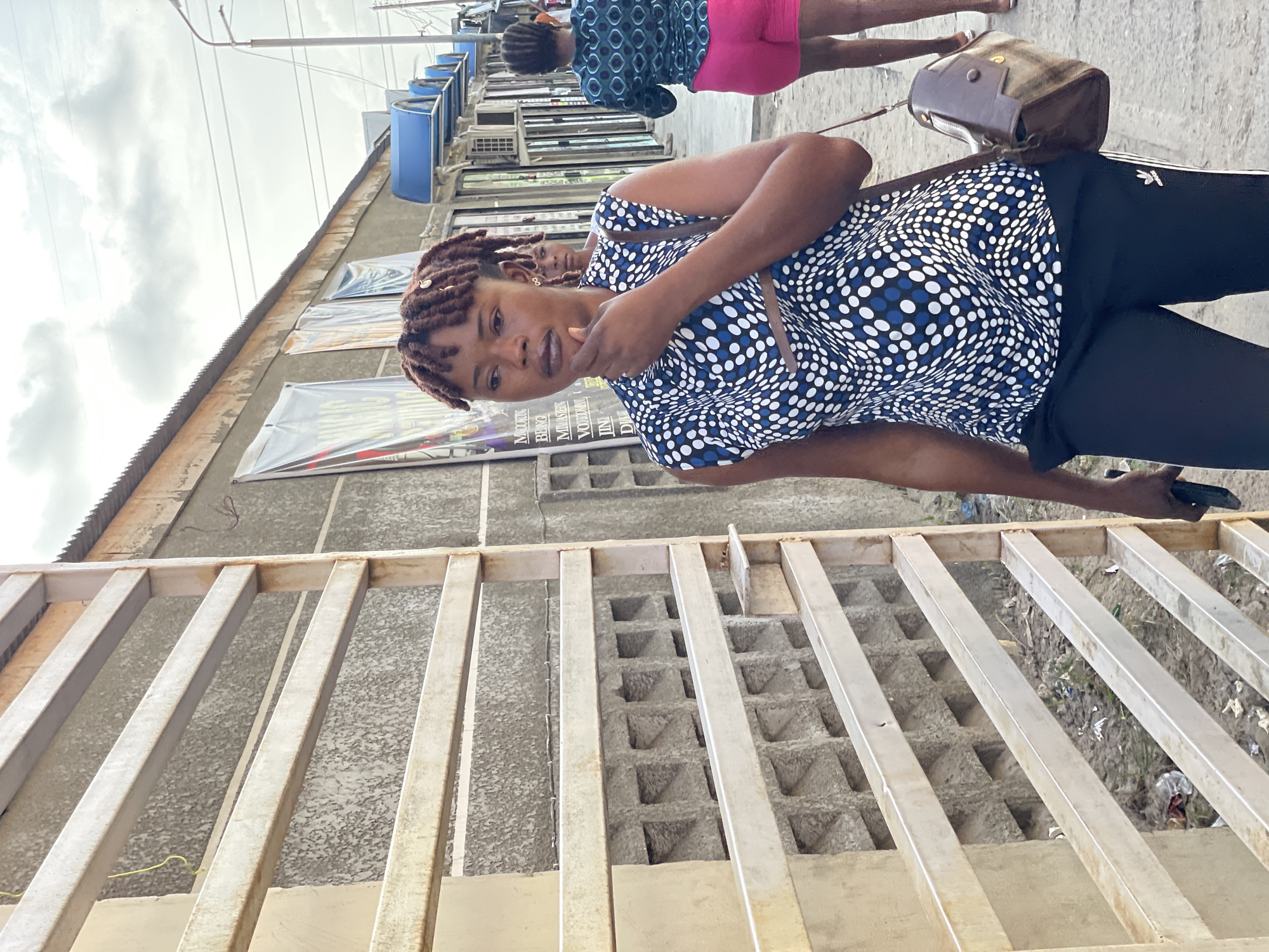 The image shows a woman with brown dreadlocks standing in front of a building in Ikorodu, Nigeria. She is wearing a patterned blue and white top and black pants. She is holding a phone in her right hand and has her left hand near her mouth, as if in thought. Behind her, another person wearing a blue patterned top and pink shorts is partially visible. The background includes a building facade with multiple storefronts, some featuring blue awnings and air conditioning units. A metal fence or railing is in the foreground. The environment appears to be an urban area with signs of commercial activity.
