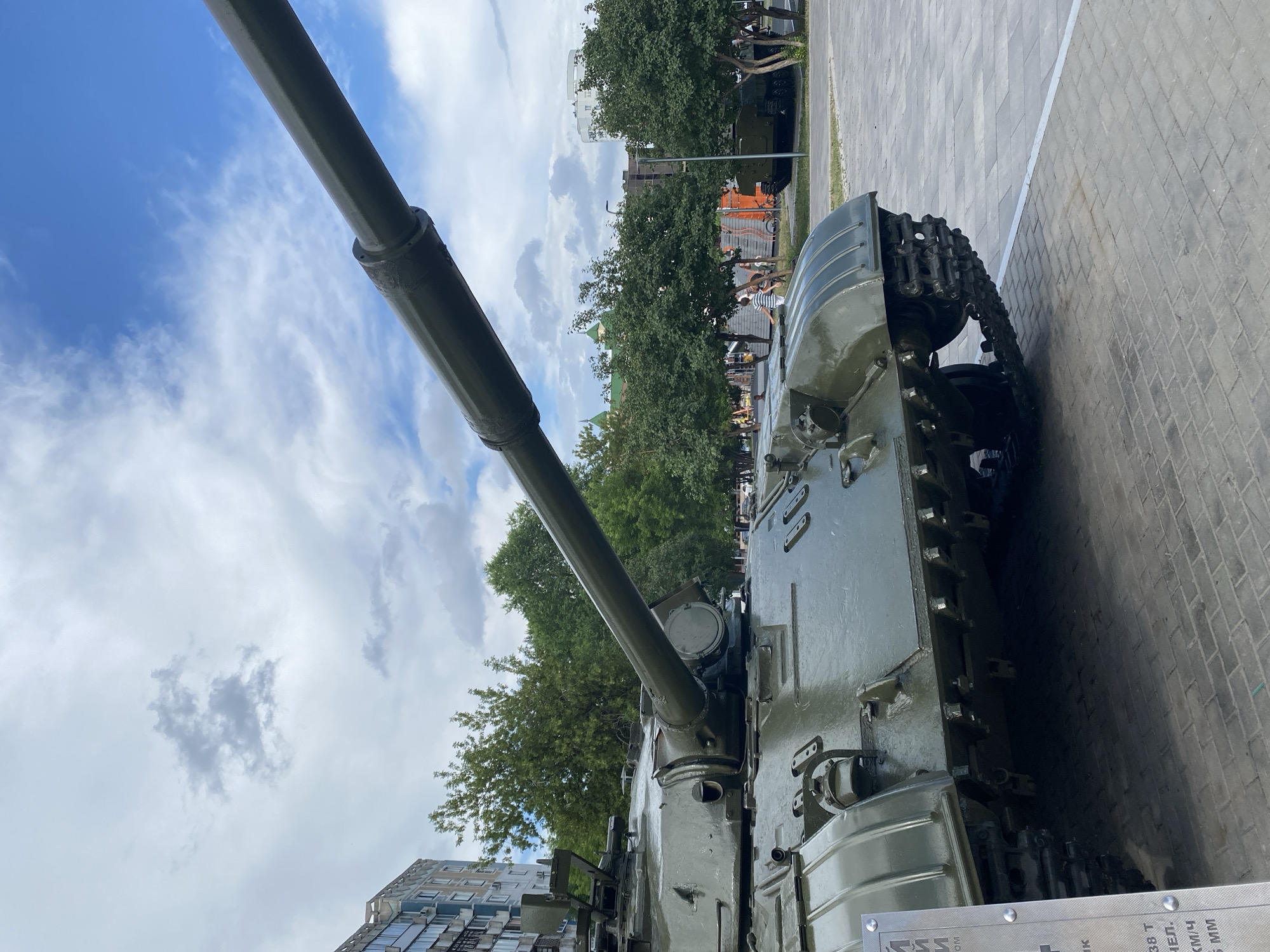 The image captures a close-up of a military tank, specifically its turret and the front part of its track, displayed outdoors on a paved area. The tank is painted in a matte olive-drab color. In the background, several trees line a sidewalk, beyond which various buildings are visible, including a multi-story residential building on the left and other structures further in the distance. The sky is partly cloudy with a mix of blue sky and white and gray clouds. The lighting suggests it is daytime. The pavement consists of rectangular blocks, and a white line demarcates an edge in the foreground. A portion of a metal information plaque with Cyrillic text is visible on the left side of the frame. Further in the background, other military vehicles and a few people can be seen, suggesting this might be a public display or monument. The image is taken from a low angle, emphasizing the size and presence of the tank.