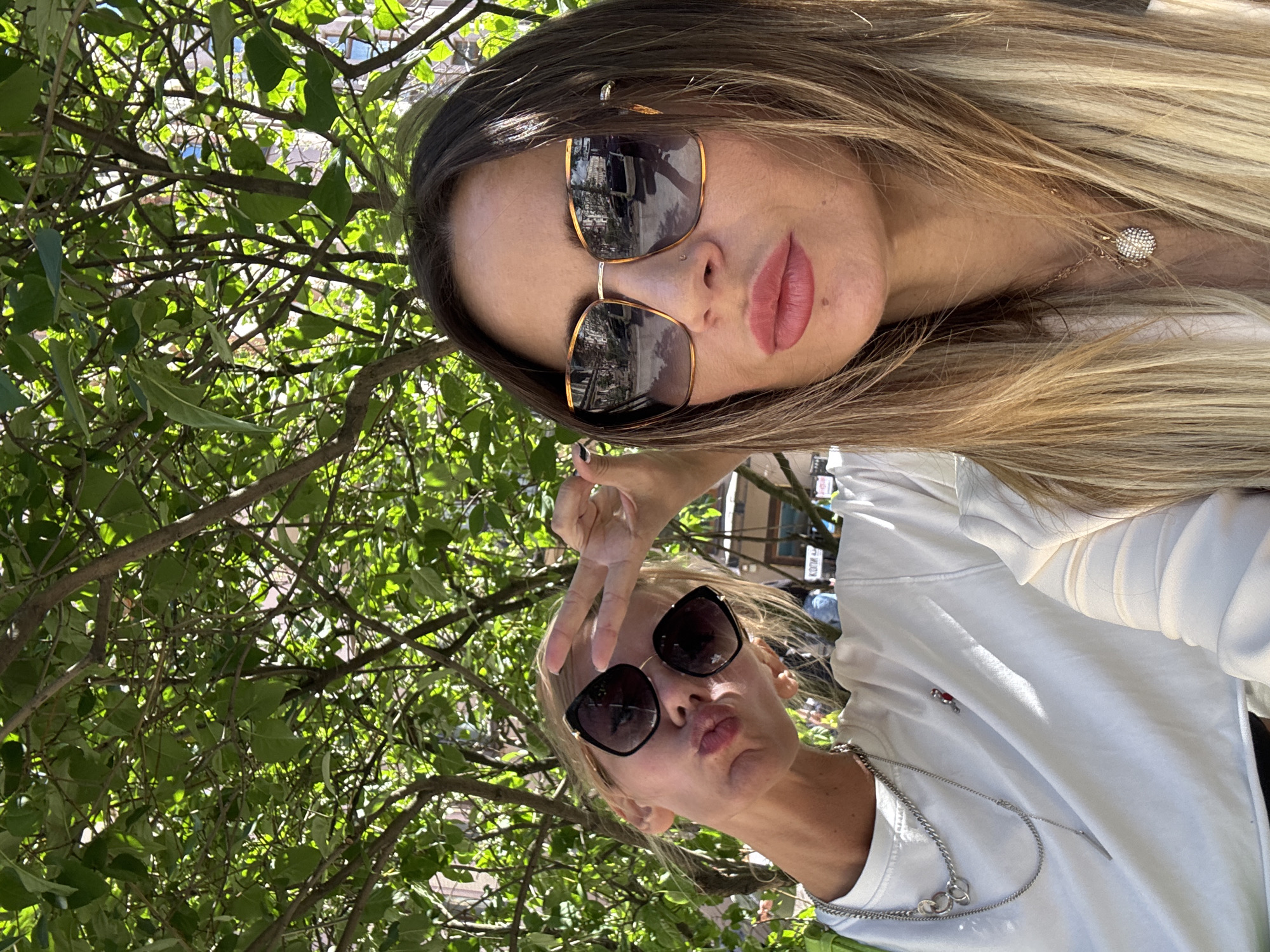 This media file captures a selfie of two young women outdoors, framed by lush green foliage from trees overhead. The image appears to be taken during daylight hours, with bright sunlight filtering through the leaves, suggesting fair weather. The women are casually dressed, with one sporting a white hoodie and the other a white top, both wearing necklaces. Both are wearing sunglasses, which obscure their eyes. One woman is making a peace sign with her fingers and has her lips puckered as if for a kiss, while the other is looking towards the camera with her lips slightly parted. Reflections are visible in their sunglasses, hinting at an urban environment with buildings and possibly vehicles. The overall mood is relaxed and cheerful, indicative of friends enjoying a sunny day together. The location context of Saint Petersburg, Russia, is provided but not visually evident within the image itself. No visible text is present in the media file.