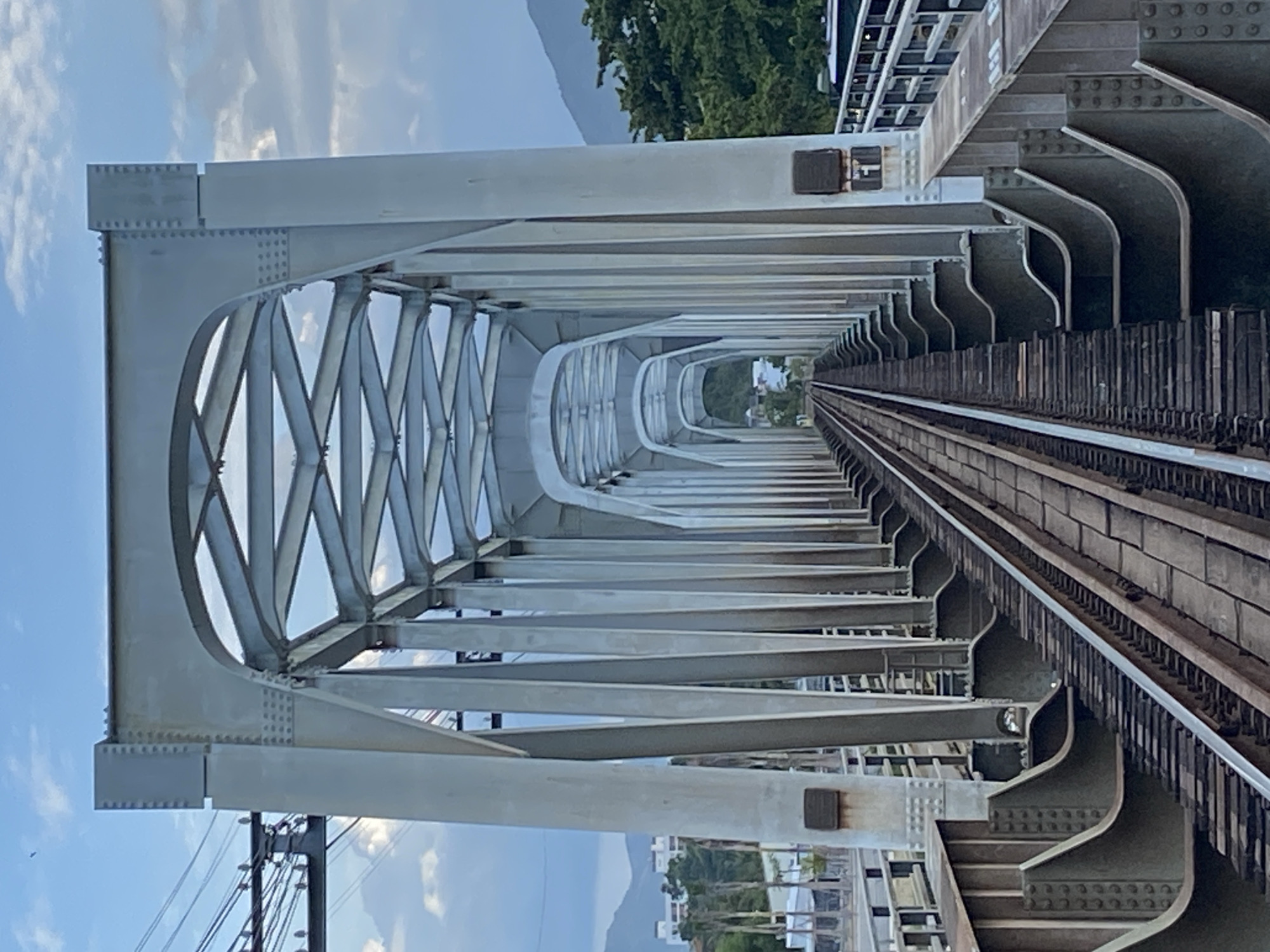 The image captures a perspective looking down train tracks that extend across a large metal truss bridge. The bridge is constructed with multiple arched supports and intricate latticework, painted in a neutral, light gray color. The train tracks are clearly visible, made of wooden ties and steel rails, and appear to be well-maintained.
On either side of the bridge, the environment shows signs of a city. To the left, there are buildings and vegetation, with utility poles and wires visible against a sky with scattered clouds. To the right, more trees and a glimpse of what appears to be a body of water or a coastal area can be seen, with distant mountains in the background. The overall scene suggests an industrial or transportation hub within a coastal city.
While the prompt requests a description of the location in Nha Trang, Vietnam, this specific visual information is not present in the image itself. The image does not contain any identifying landmarks, signs, or specific architectural styles that definitively place it in Nha Trang, Vietnam. Therefore, while the scene depicts a bridge in a city, its precise location cannot be determined from the visual data alone.