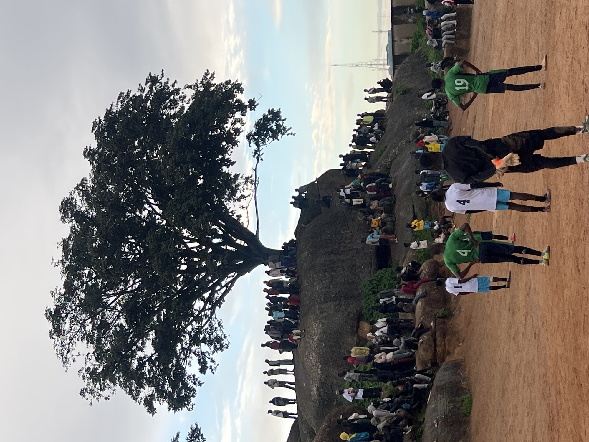 In Jos, Nigeria, a group of people are gathered on a large rocky outcrop beneath a mature tree with a wide canopy. The sky above is overcast with patches of blue peeking through. On the rock formation, numerous individuals are standing and sitting, observing something below. The ground in the foreground is a reddish-brown dirt, suggesting a dry, possibly dusty environment. In the middle ground and foreground, several young men are positioned as if participating in or watching a soccer game, with some wearing jerseys. One individual in the foreground is wearing a green jersey with the number 4 on the back, and another has a green jersey with the number 19. Another person is wearing a white jersey with the number 8. The overall atmosphere appears to be one of casual gathering and recreation. In the distance, a tall communication tower is visible, along with some buildings and what appears to be a wall or fence. The scene captures a slice of daily life in this Nigerian city, combining natural elements with human activity.